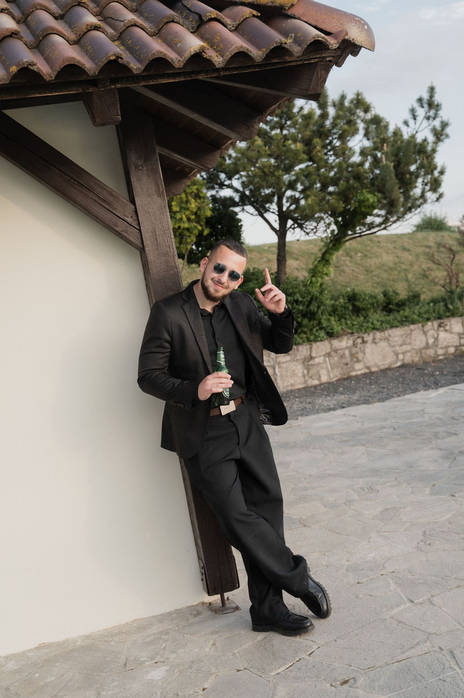 Stylish wedding guest in black suit leaning against wall holding champagne bottle and glass outdoors.