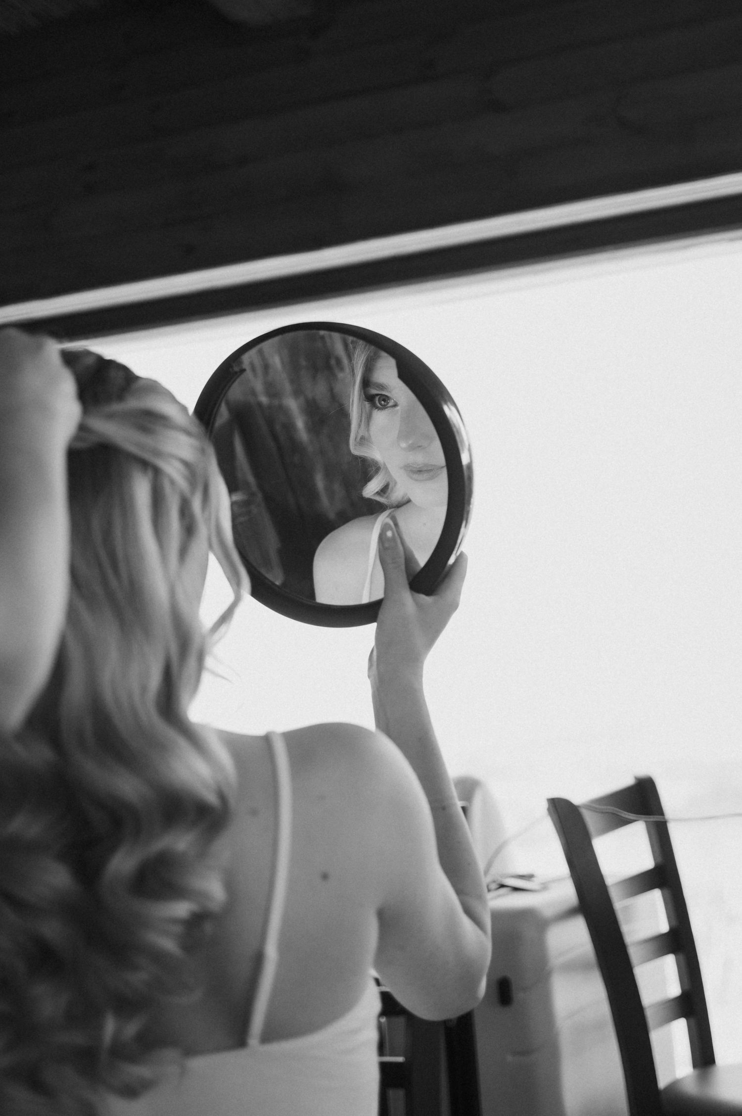 Blonde bride applying lipstick while gazing into a round handheld mirror in a black and white wedding photo.