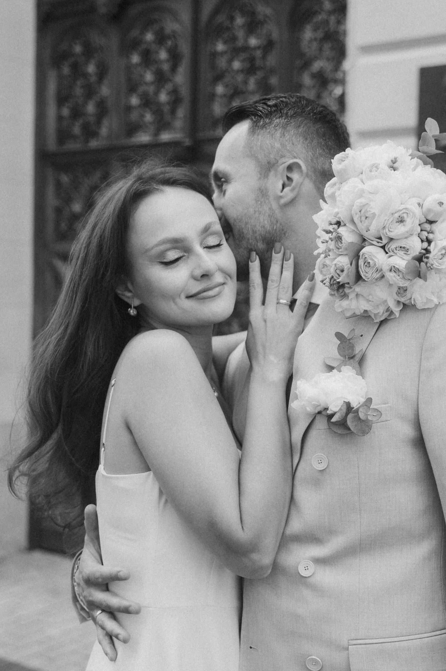 Romantic wedding couple embraces in front of ornate architectural details.