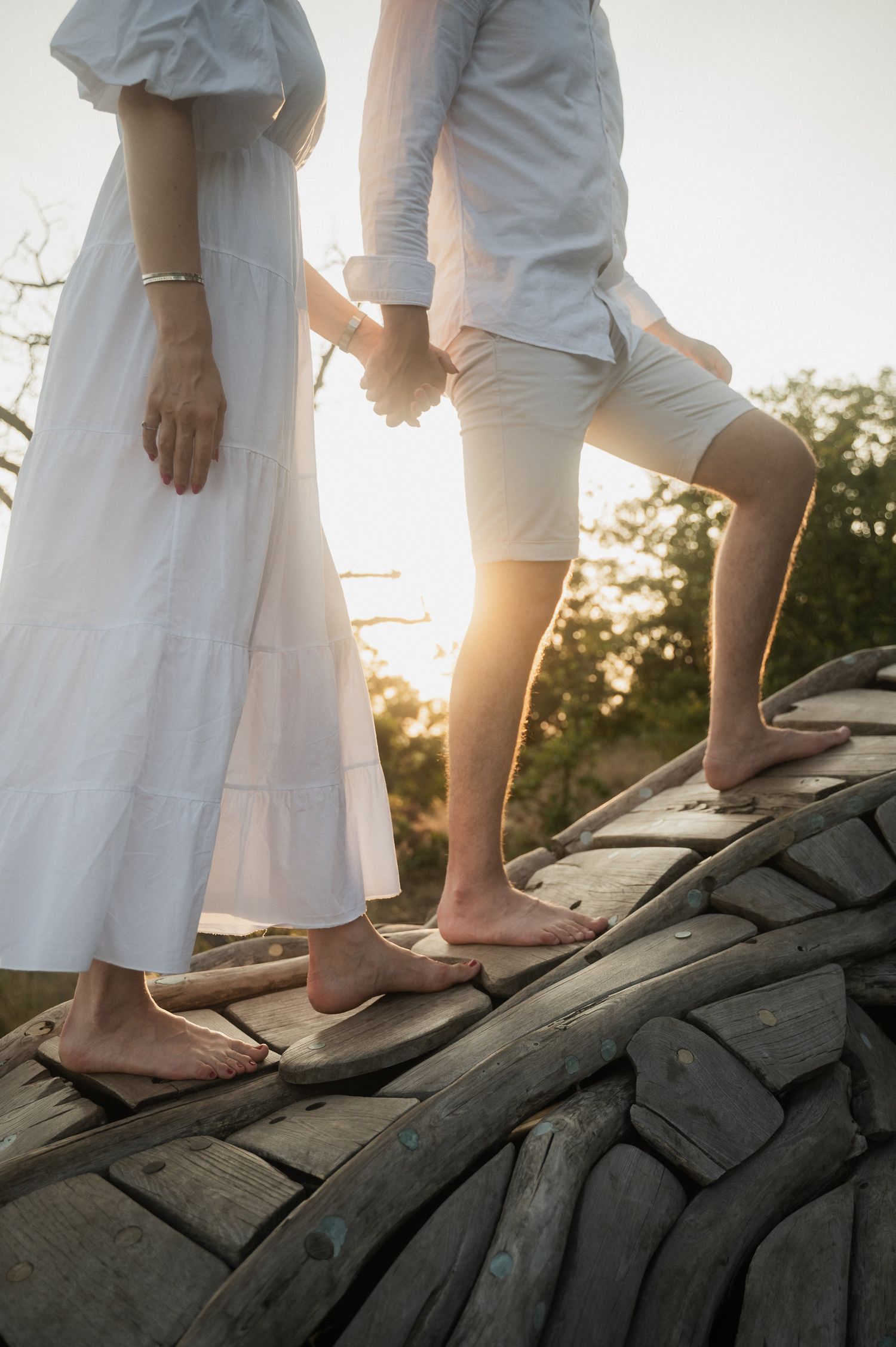 A couple holding hands while standing on large rocks at sunset, wearing white and beige summer clothing.