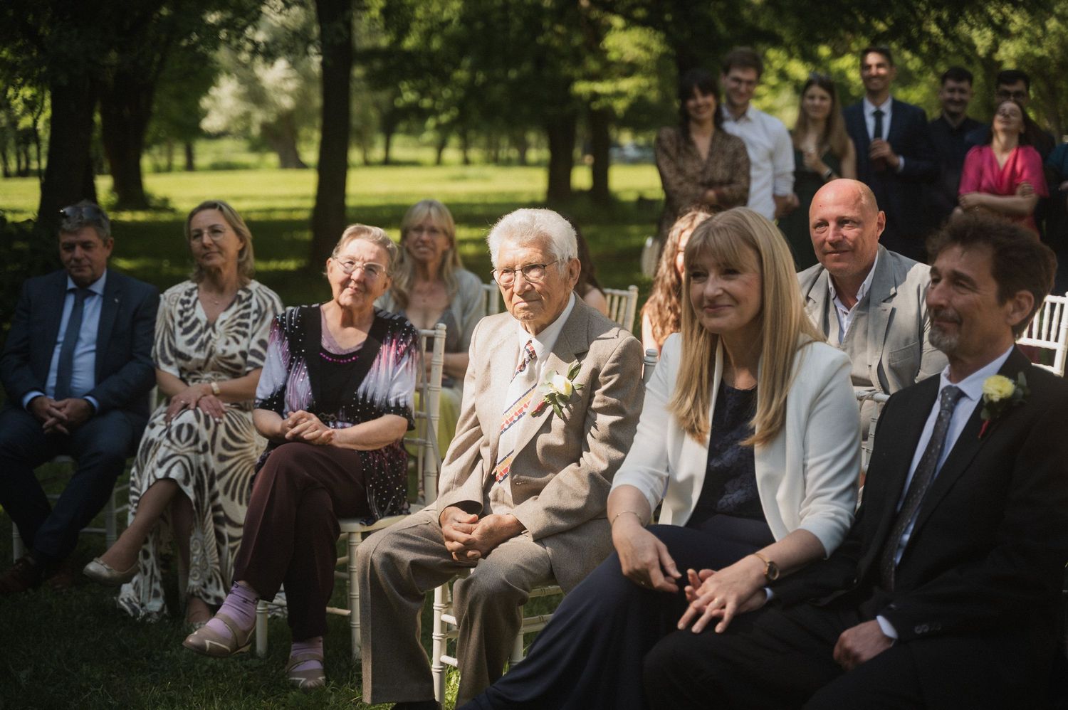 Wedding guests seated in rows at an outdoor garden ceremony smile while watching the proceedings on a sunny day.