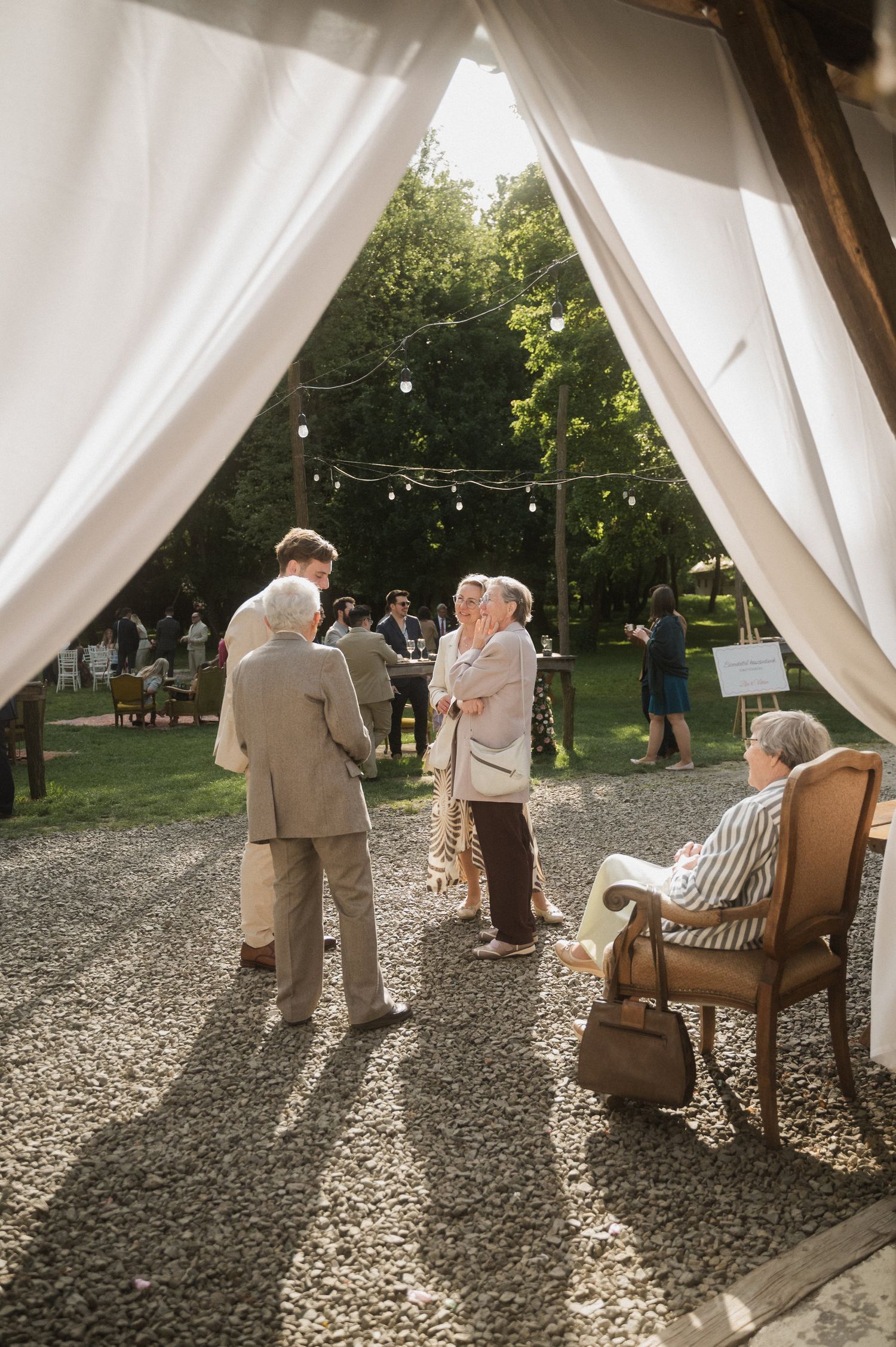 A couple sits under a white draped canopy in an outdoor garden setting during a photoshoot.