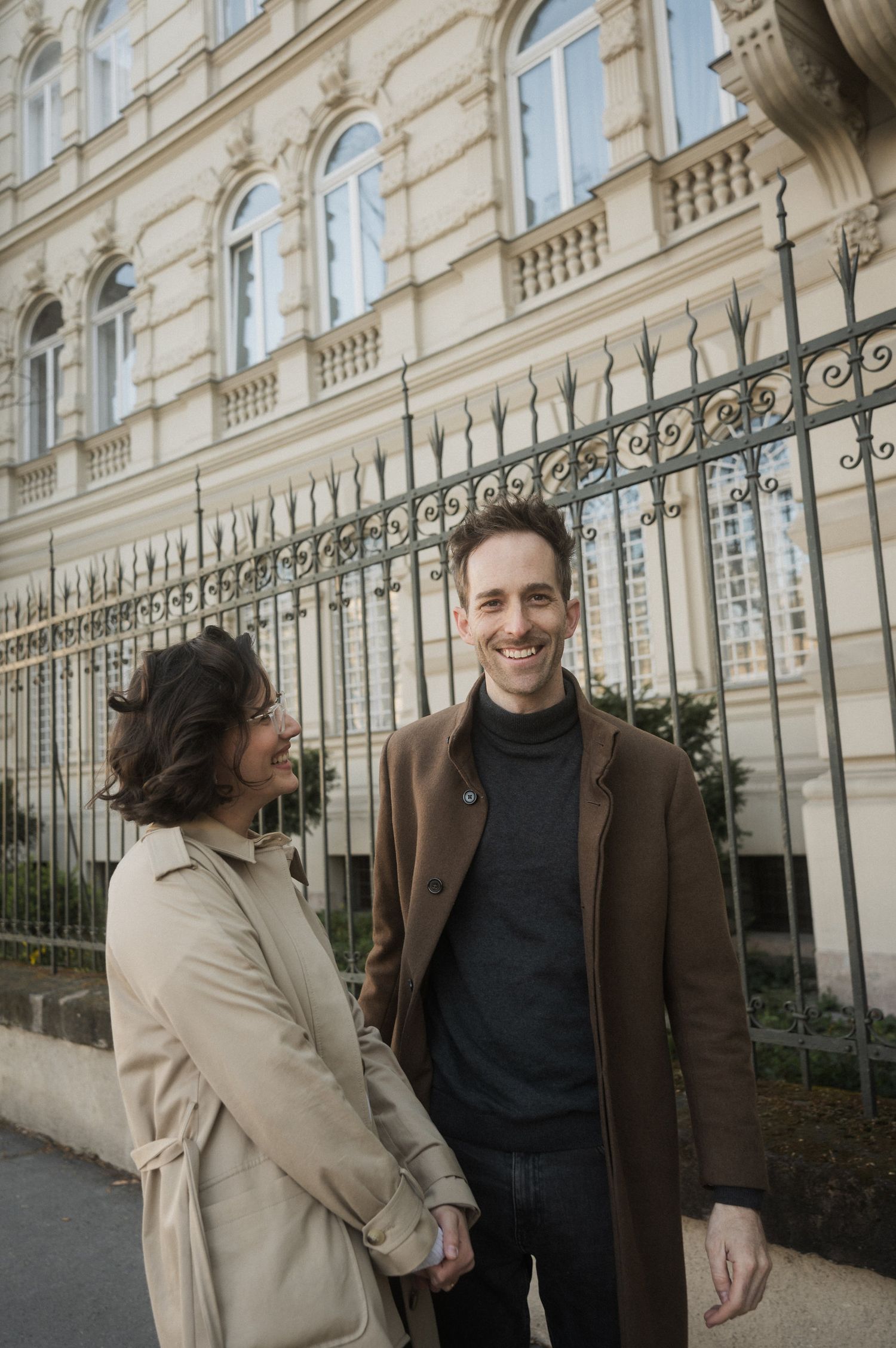 A couple shares a candid moment while walking together outside an ornate historic building with wrought iron fencing.