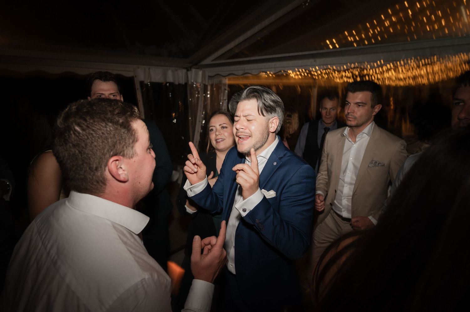 Guests celebrating and dancing energetically at a wedding reception under warm string lights.