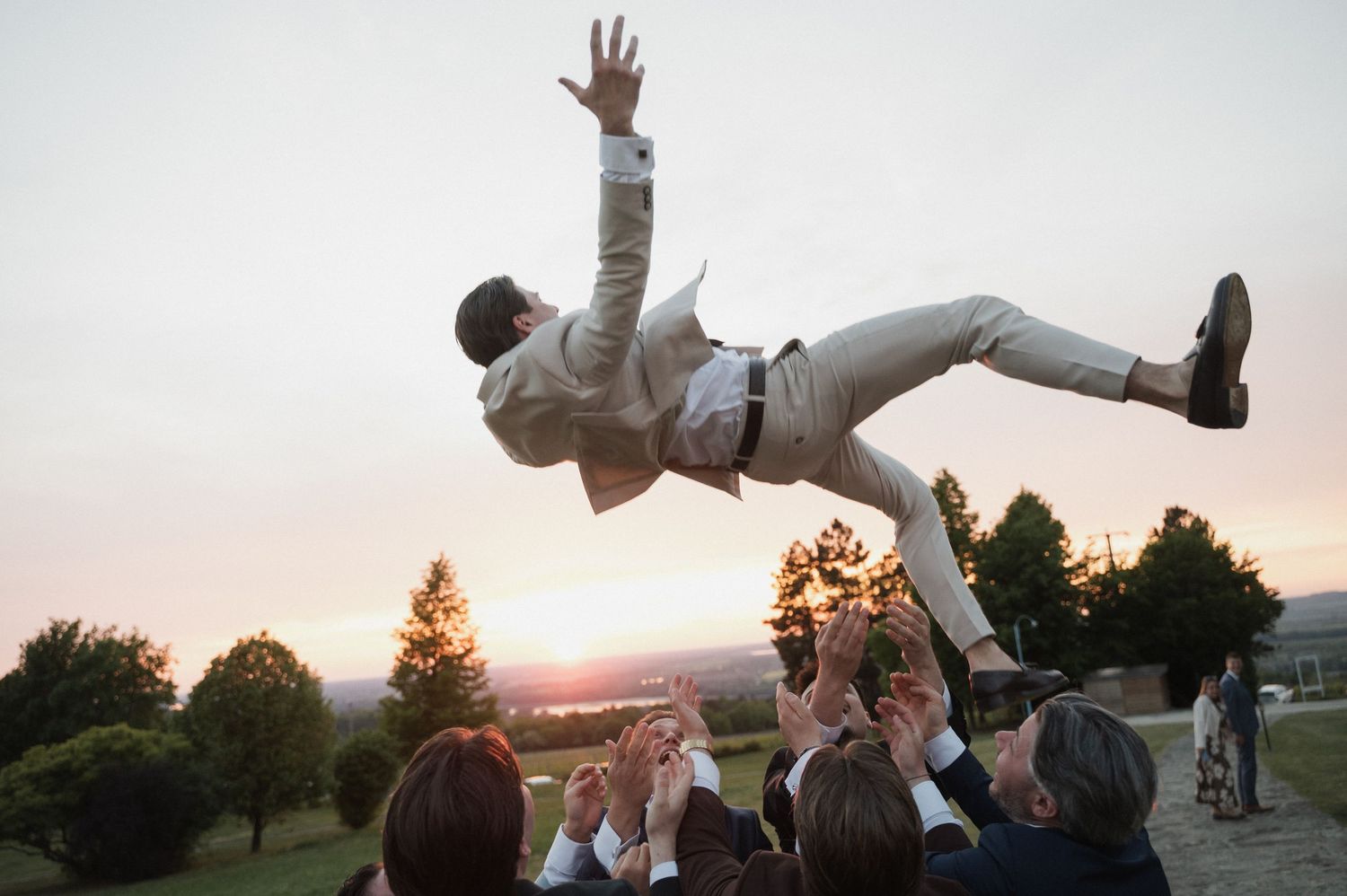 Groomsmen tossing groom in the air at outdoor wedding celebration during golden hour sunset.