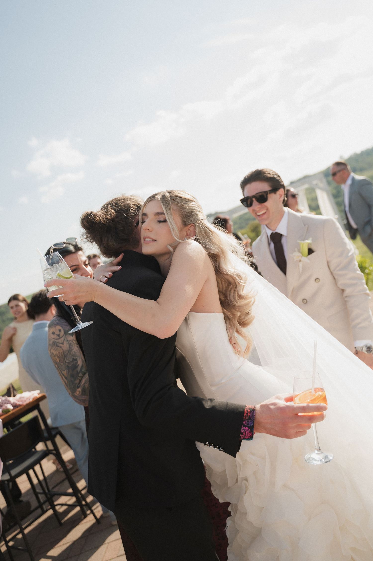 Bride in white gown hugging guest in black dress while holding champagne at outdoor reception.