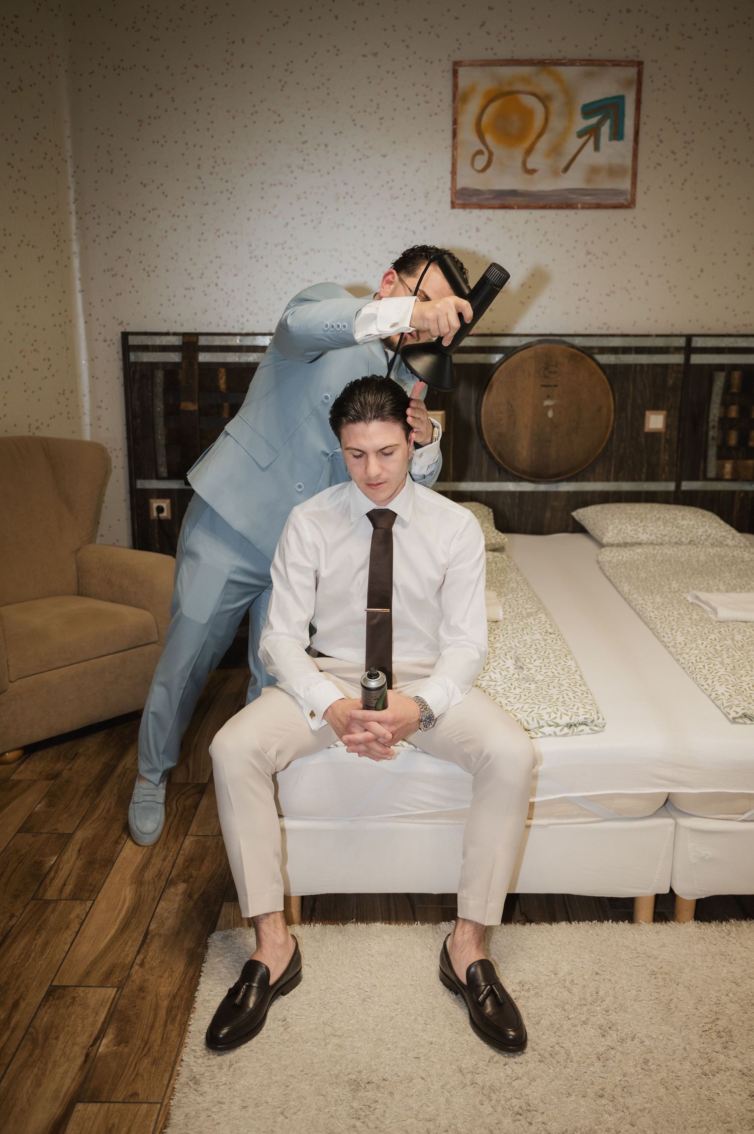 Groom sits on bed while groomsman styles his hair with a blow dryer before the wedding ceremony.