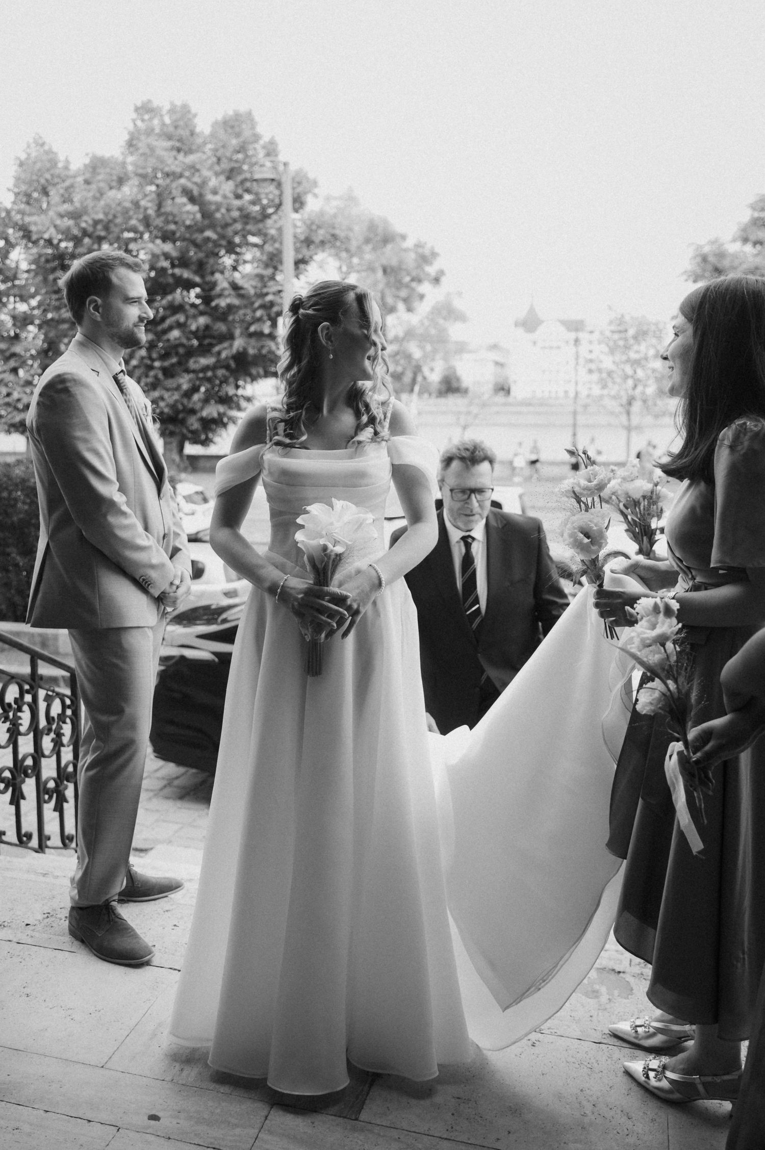 Bride in elegant white gown holds bouquet during outdoor wedding ceremony while bridesmaid adjusts her train.