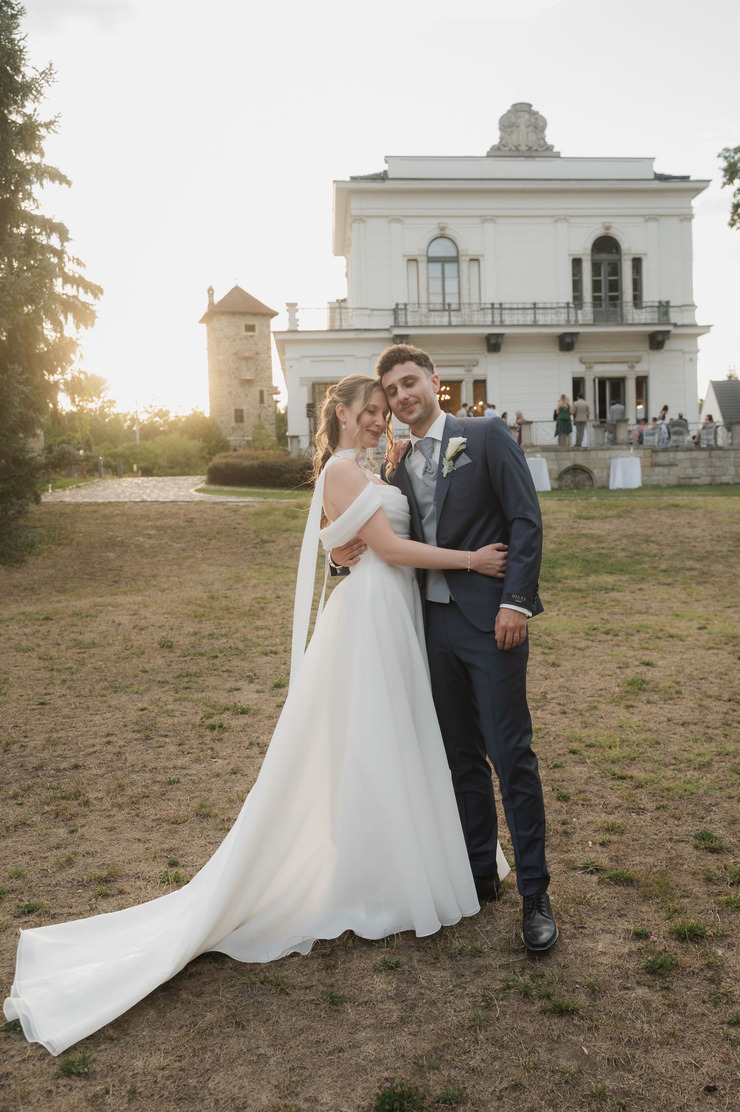 Bride and groom pose at golden hour outside a grand white villa, bride wearing a flowing white cape wedding gown.