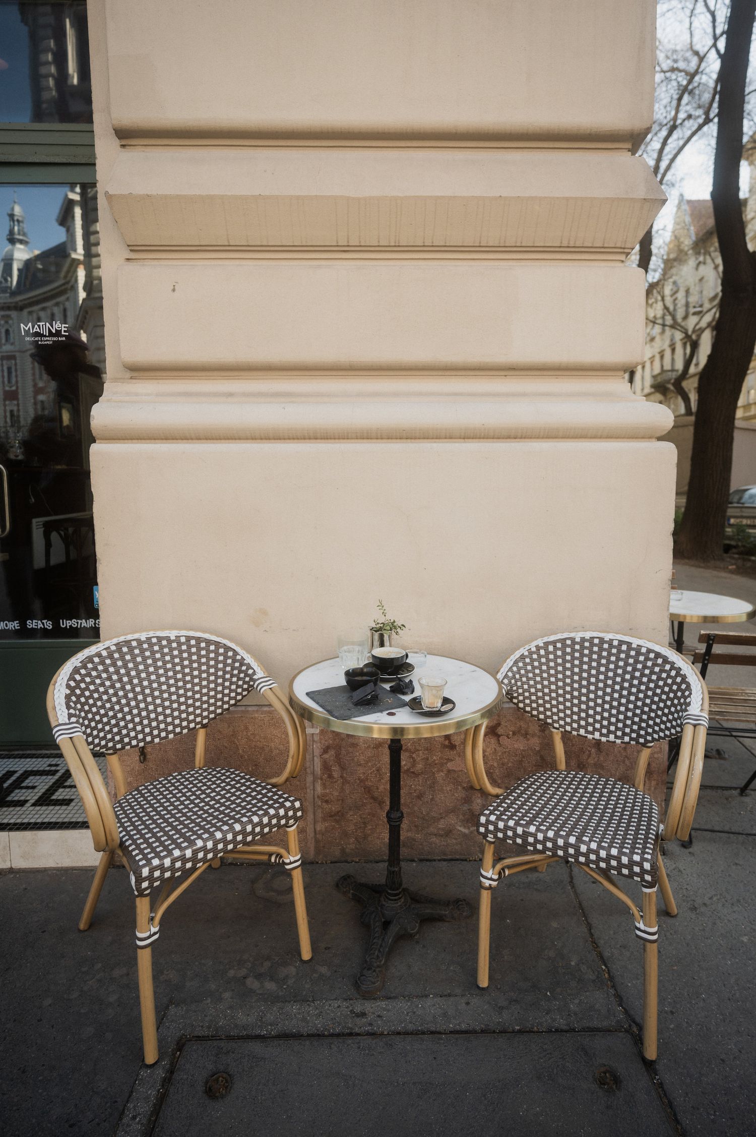 Two black and white rattan bistro chairs with wooden frames sit empty against a beige wall on a sidewalk.