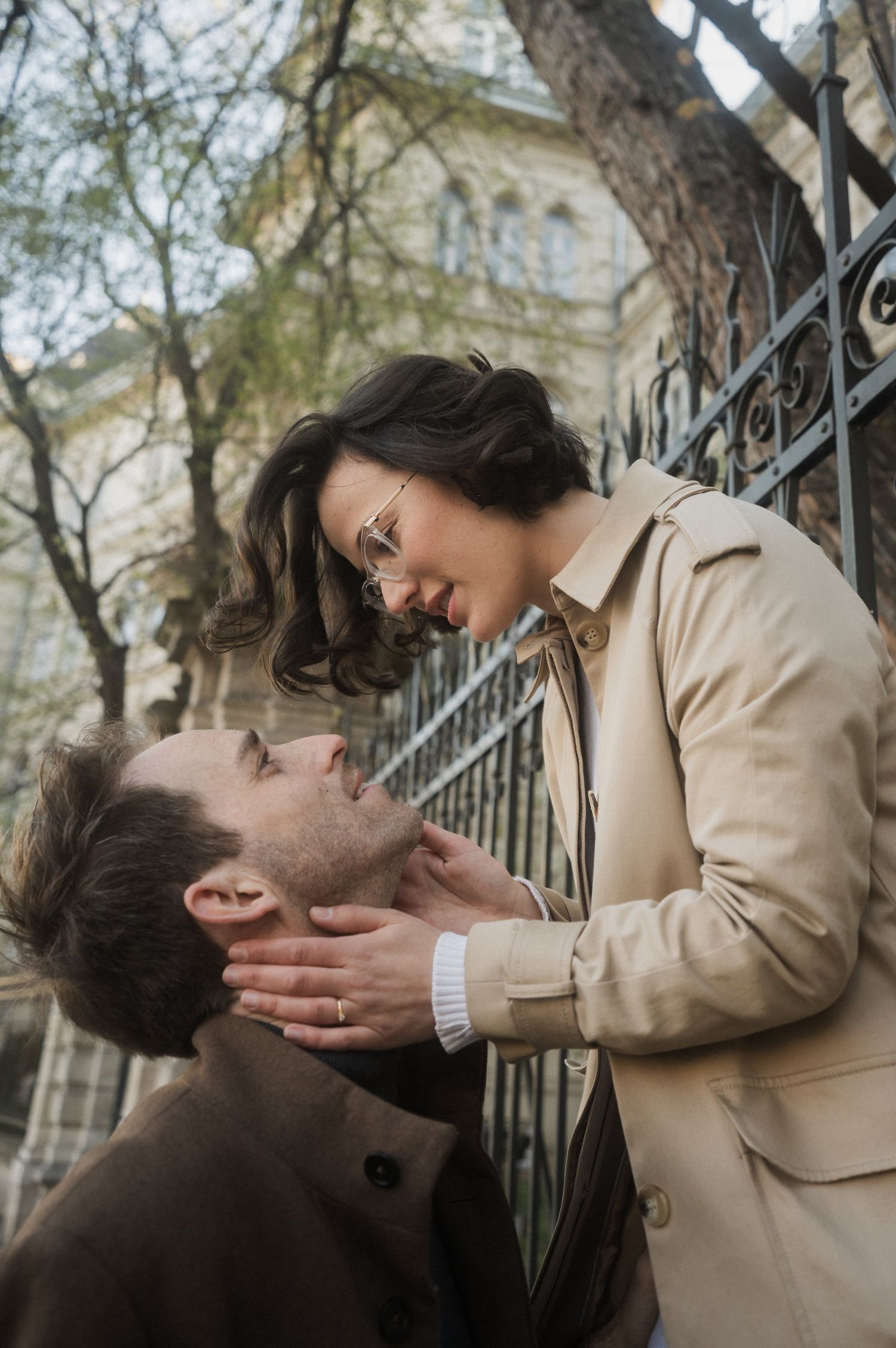 A romantic couple shares an intimate moment against an iron gate with a building facade visible in the background.