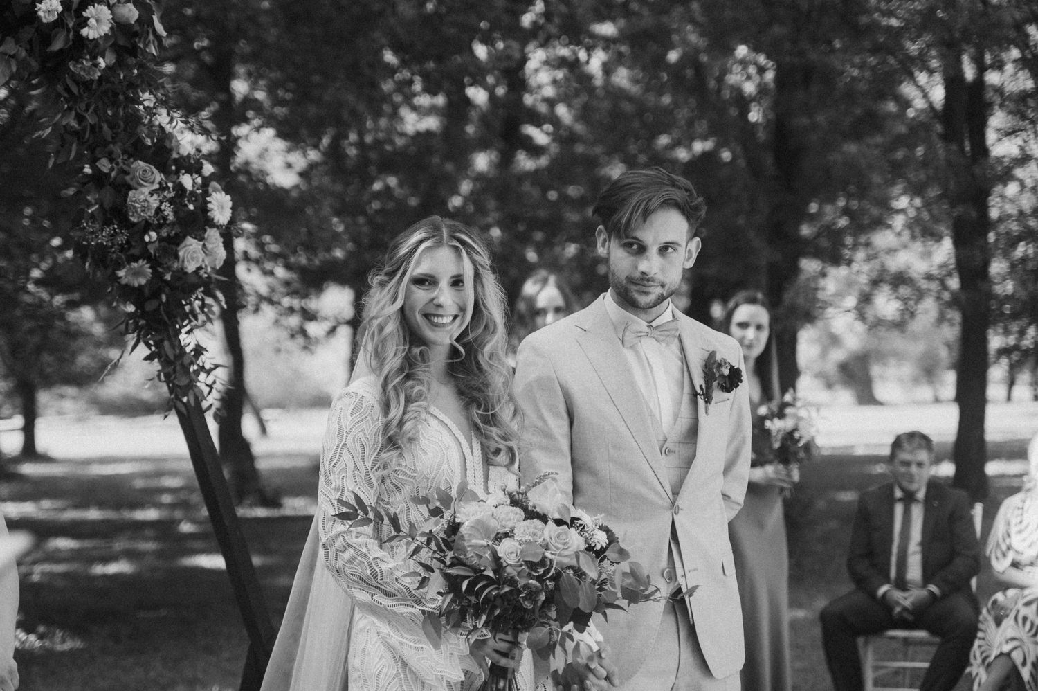 Black and white photo of a bride and groom at an outdoor wedding ceremony with trees in the background.