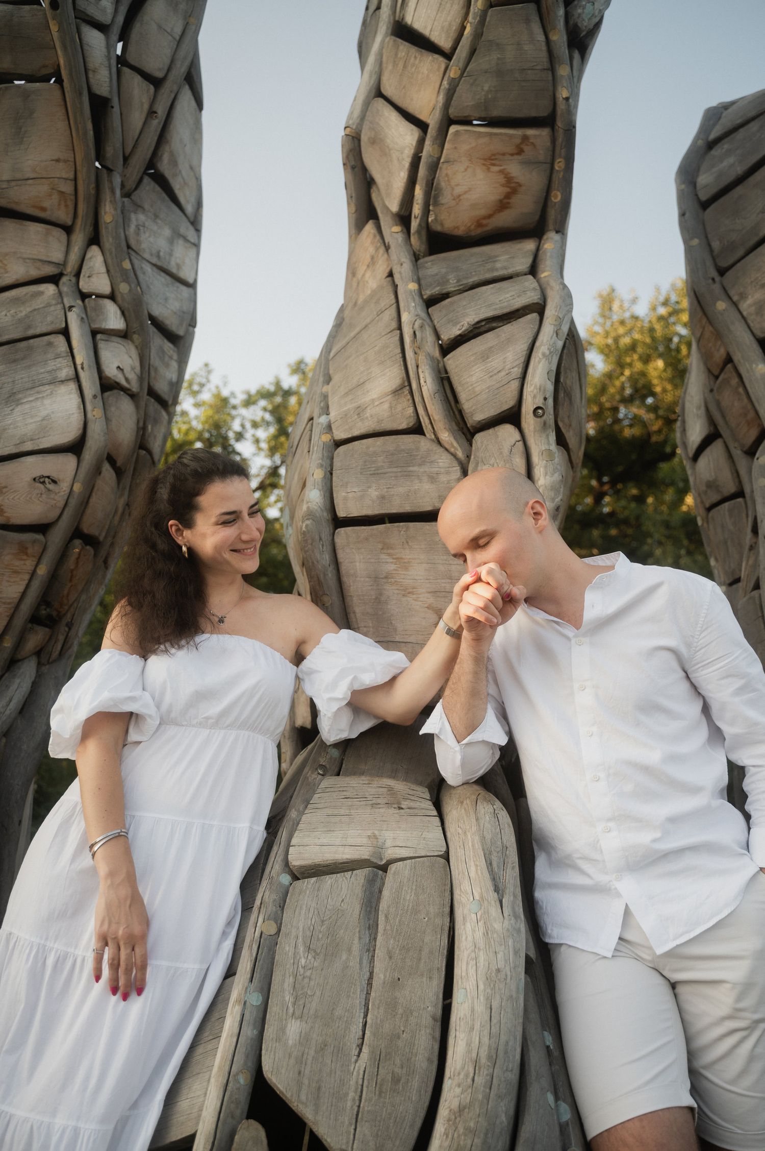 A romantic couple in white outfits poses against large wooden sculptures in an outdoor setting during golden hour.