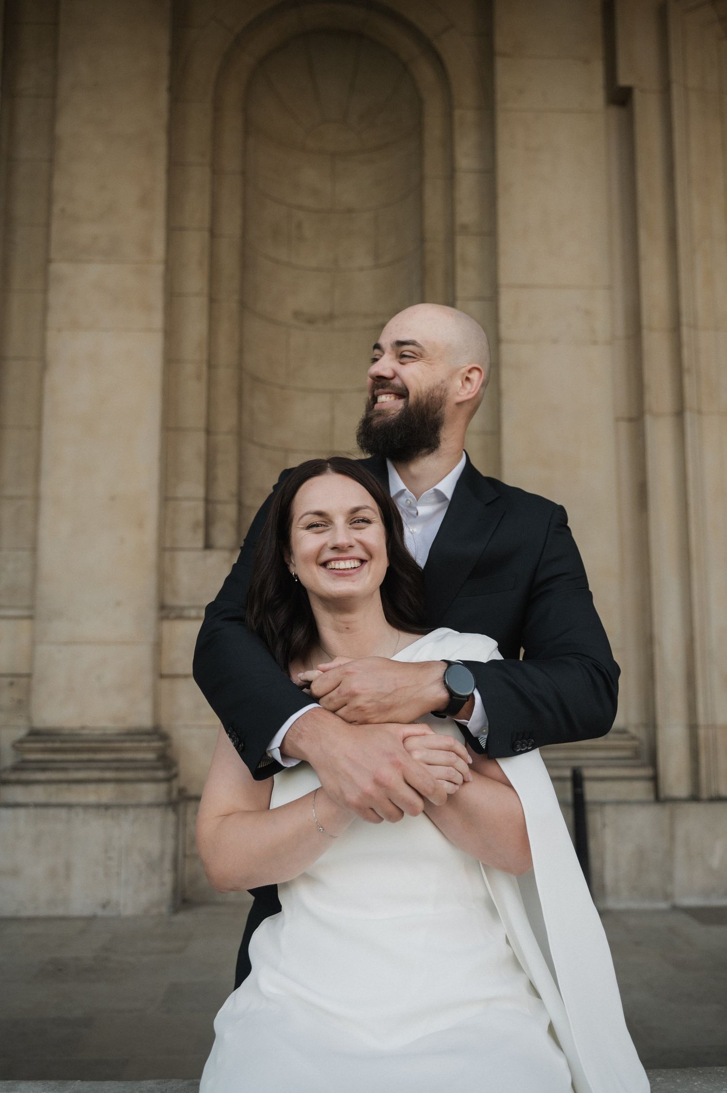 A romantic wedding couple embraces and laughs together under stone archways.