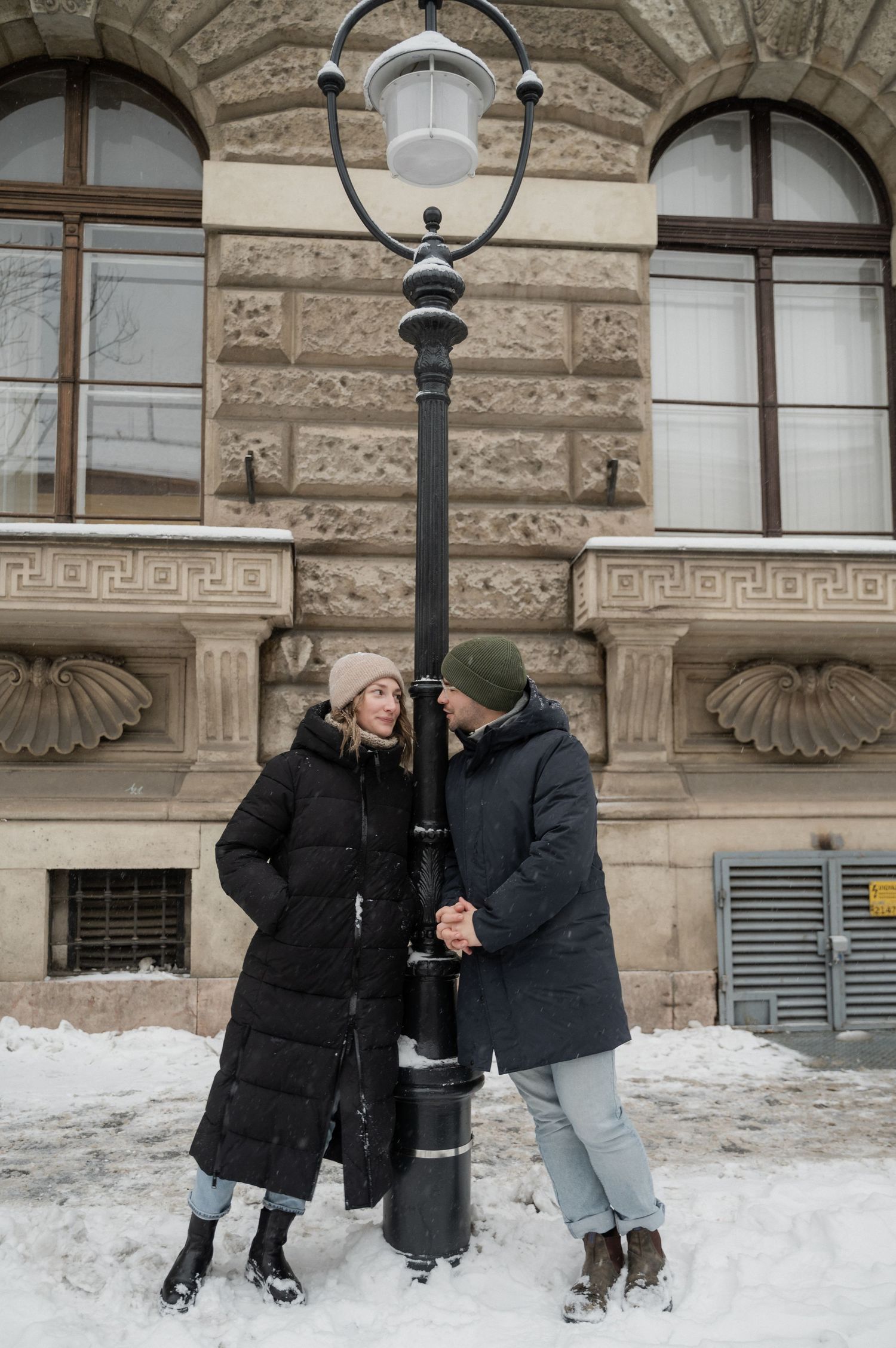 Two people in winter coats stand close together by an ornate lamppost in the snow.