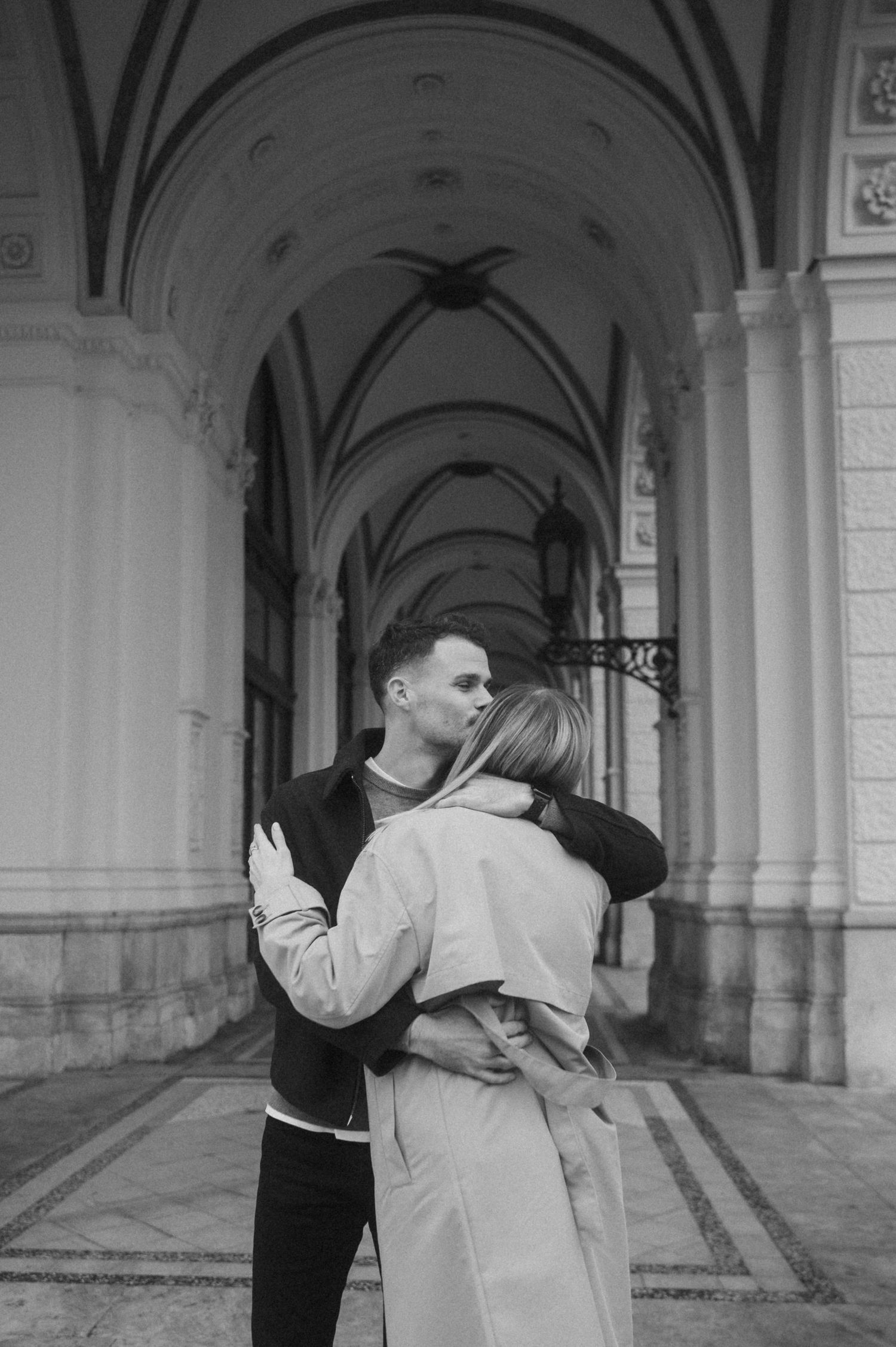 A couple shares a playful moment under ornate arched corridors in a vintage black and white photograph.