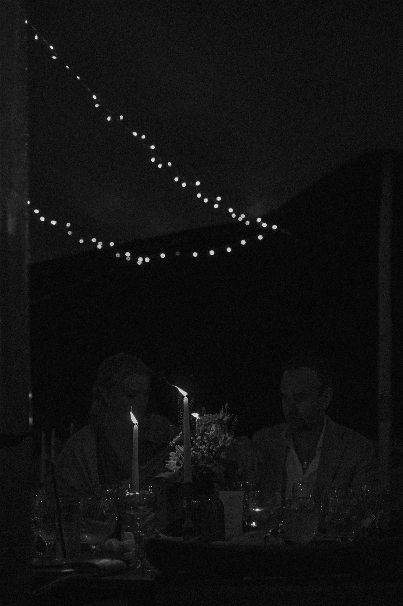 String lights illuminate a dark evening reception with silhouetted figures and candlelight.