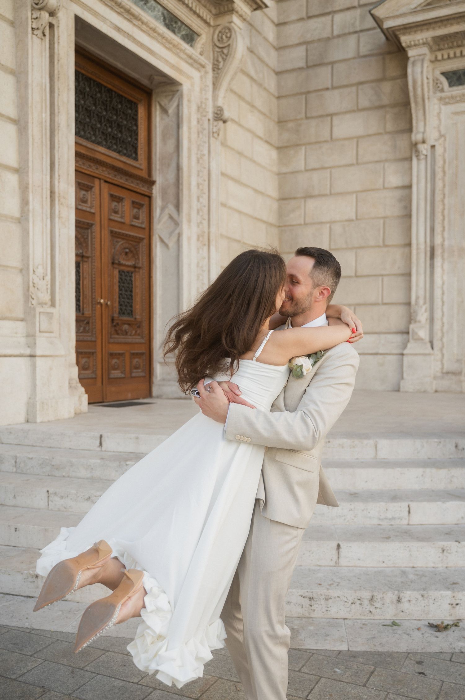 Groom in light suit spins bride in white dress on stone courtyard.