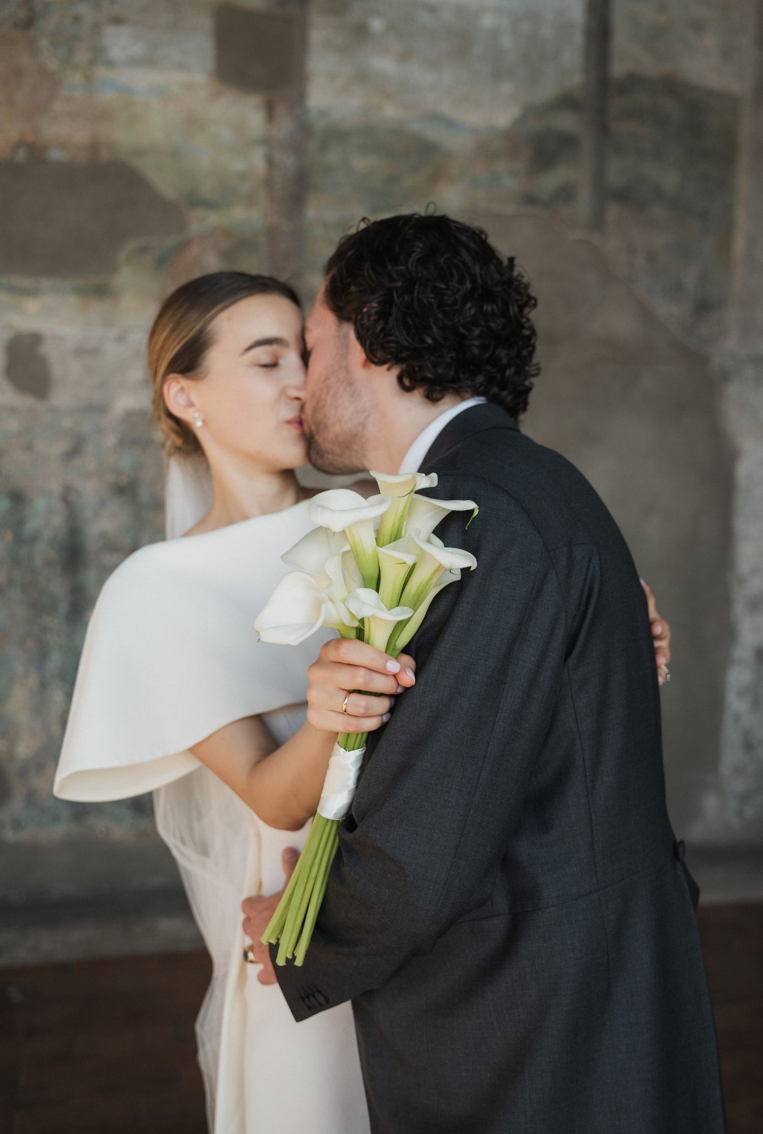 Intimate wedding portrait with white calla lily bouquet against textured stone walls.
