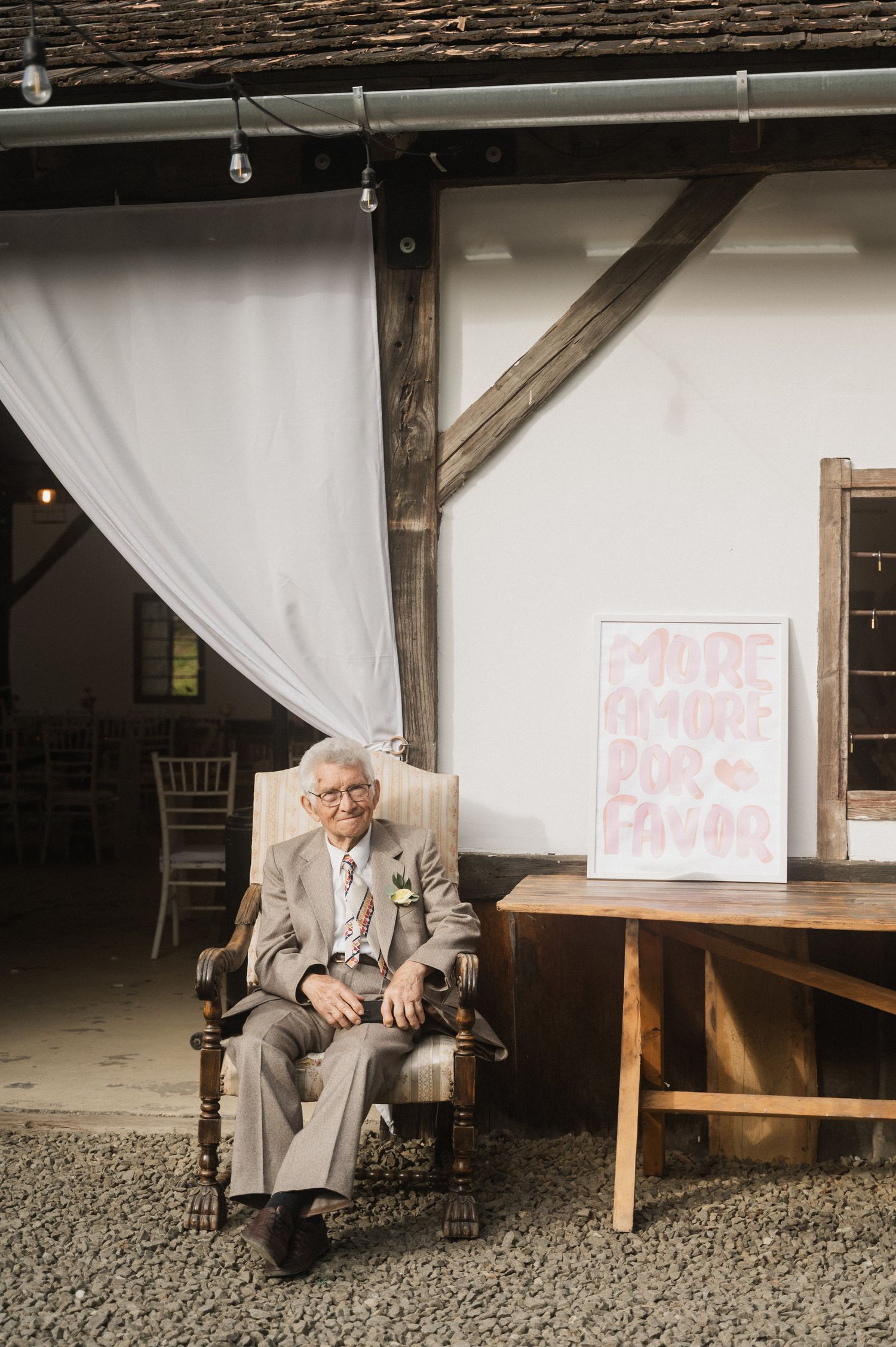 A person in a light gray suit sits on a wooden chair against a white backdrop at an outdoor venue.