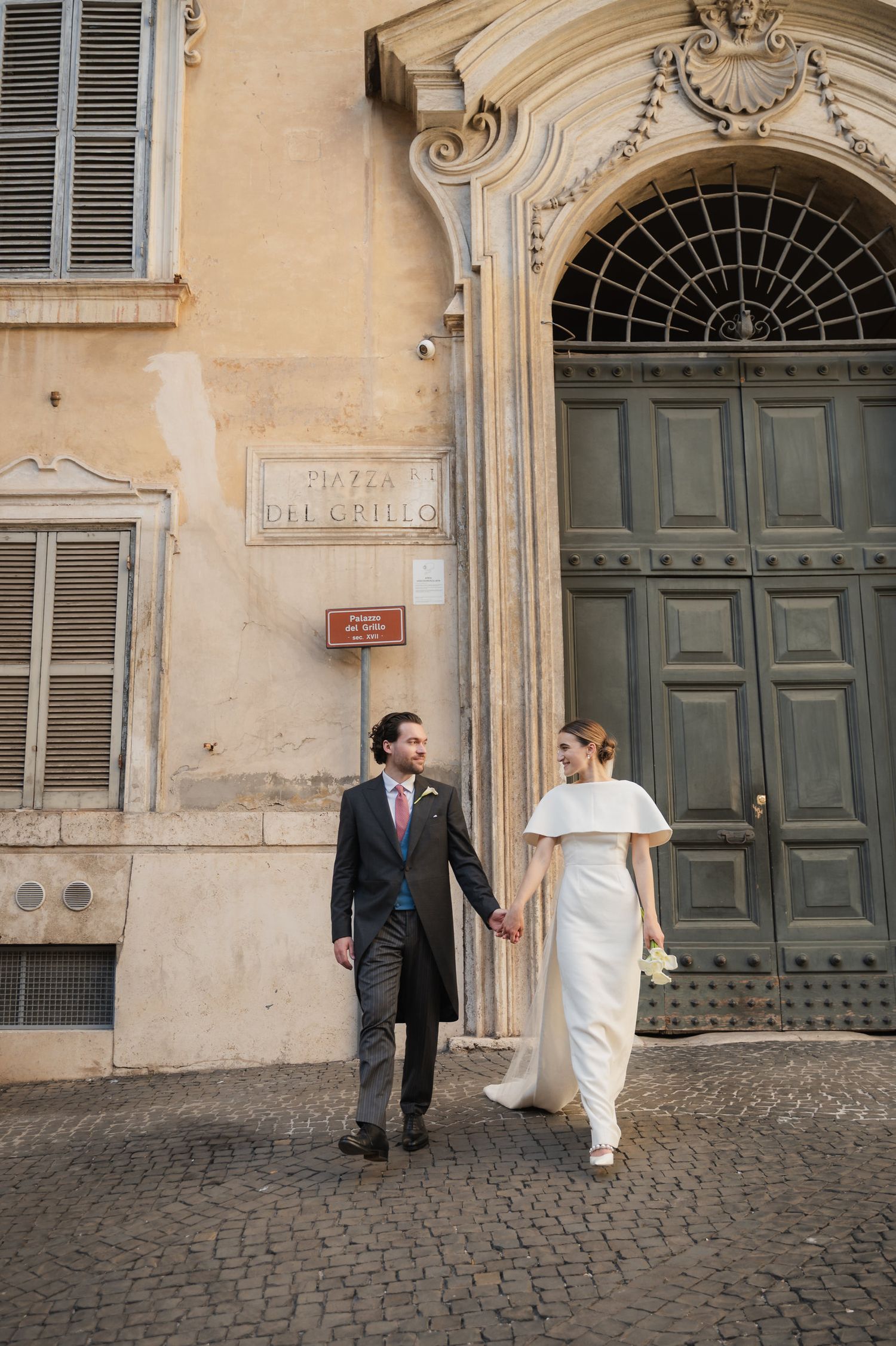 Wedding couple walks along cobblestone street past ornate green doors and beige building facade.