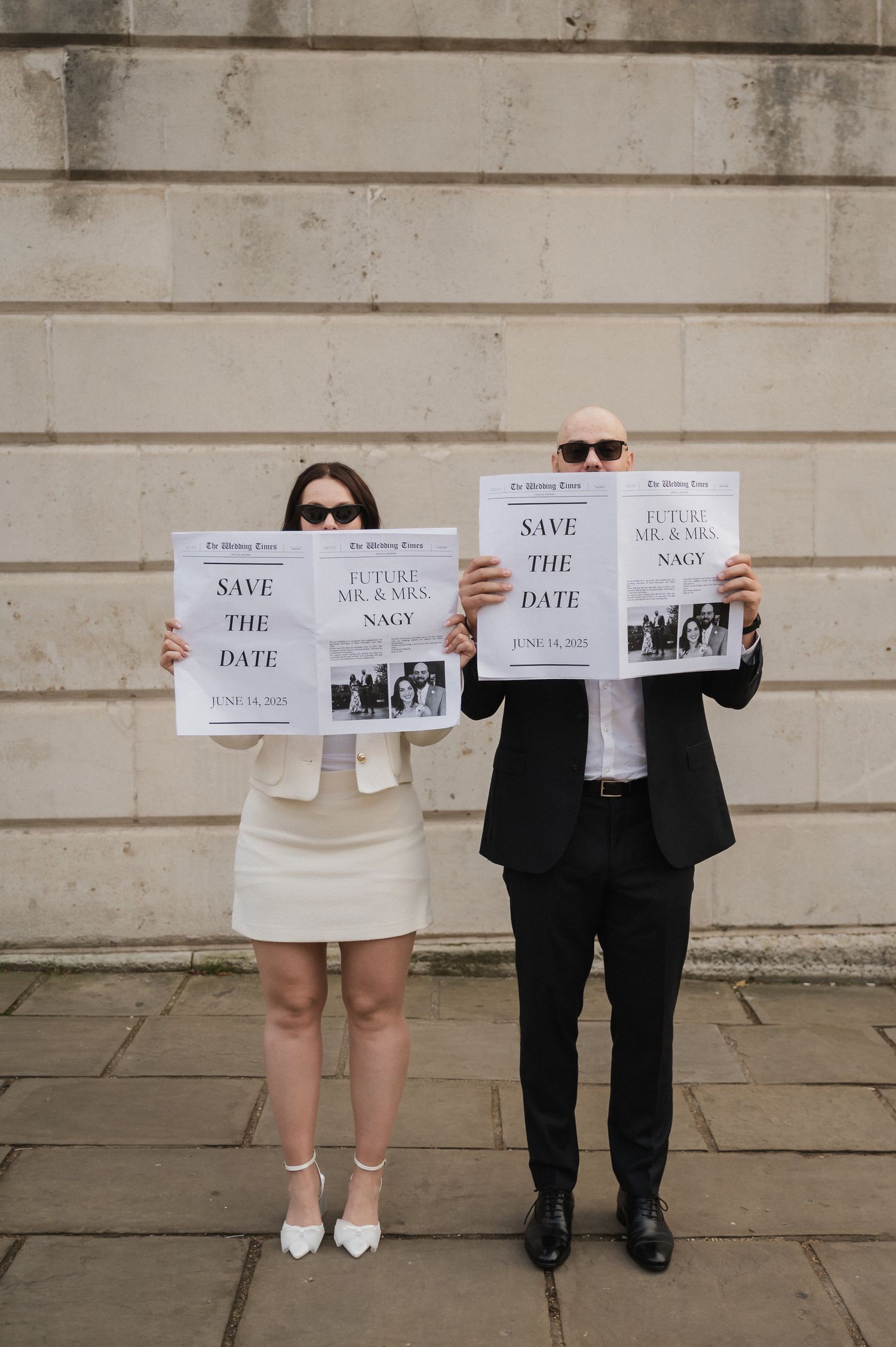Two people hold up newspaper-style save the date announcements against a stone wall.