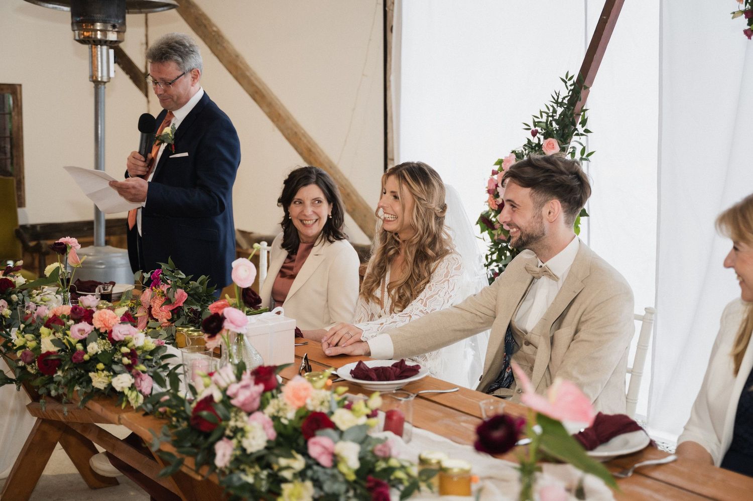 Wedding party seated at a rustic wooden table decorated with pink and white floral arrangements during reception.