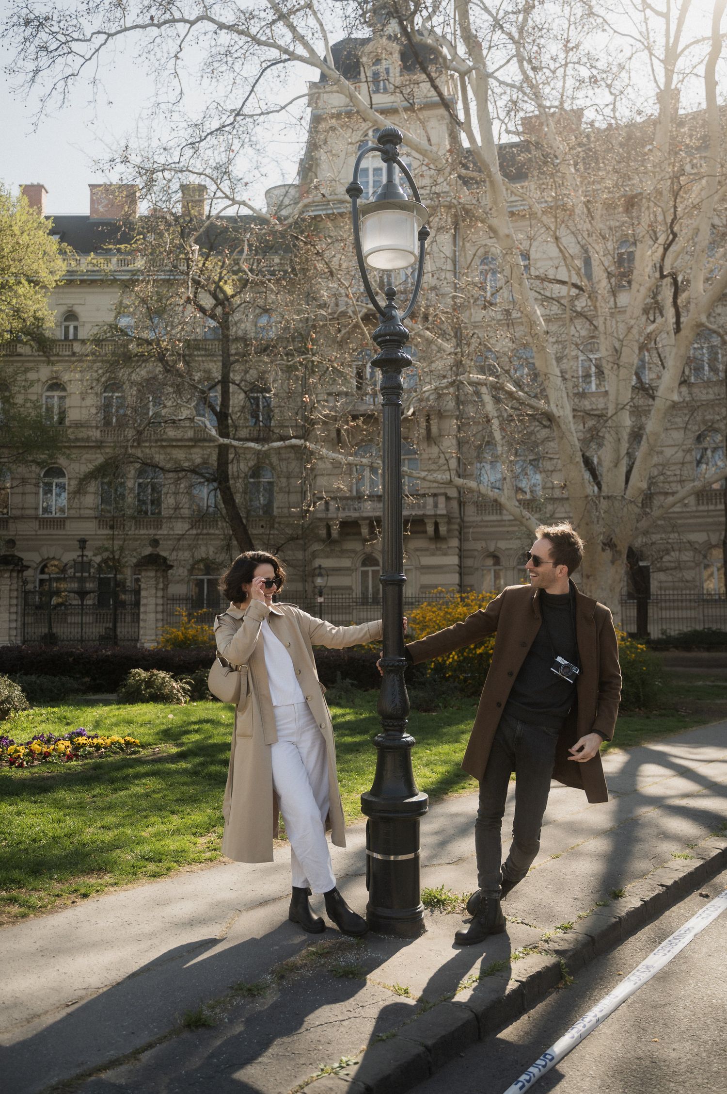 Two people stroll along a tree-lined walkway beside classic architecture as sunlight streams through bare branches.