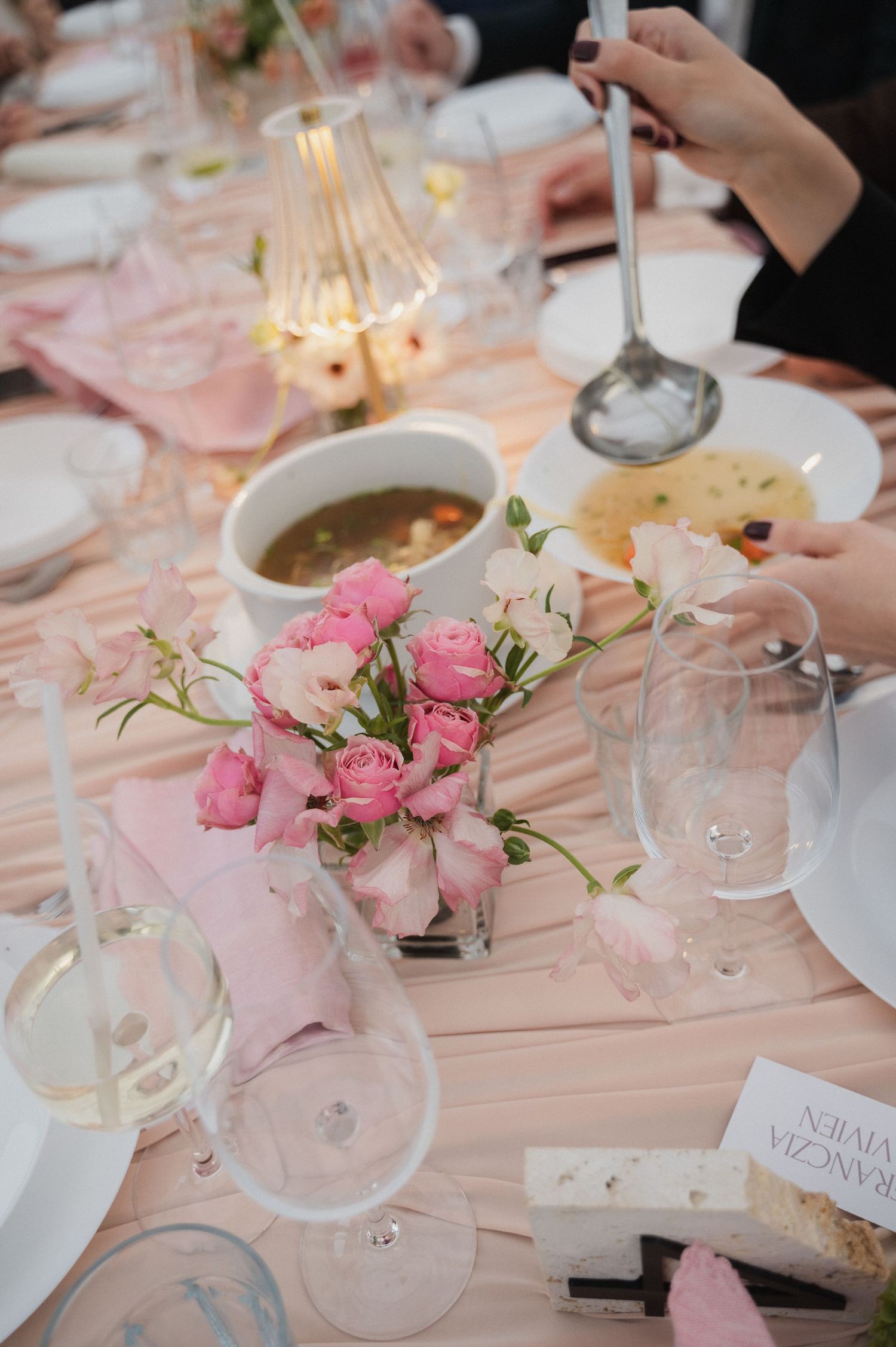 Elegant wedding reception table with pink floral centerpiece, wine glasses, and soup bowls on a blush tablecloth.