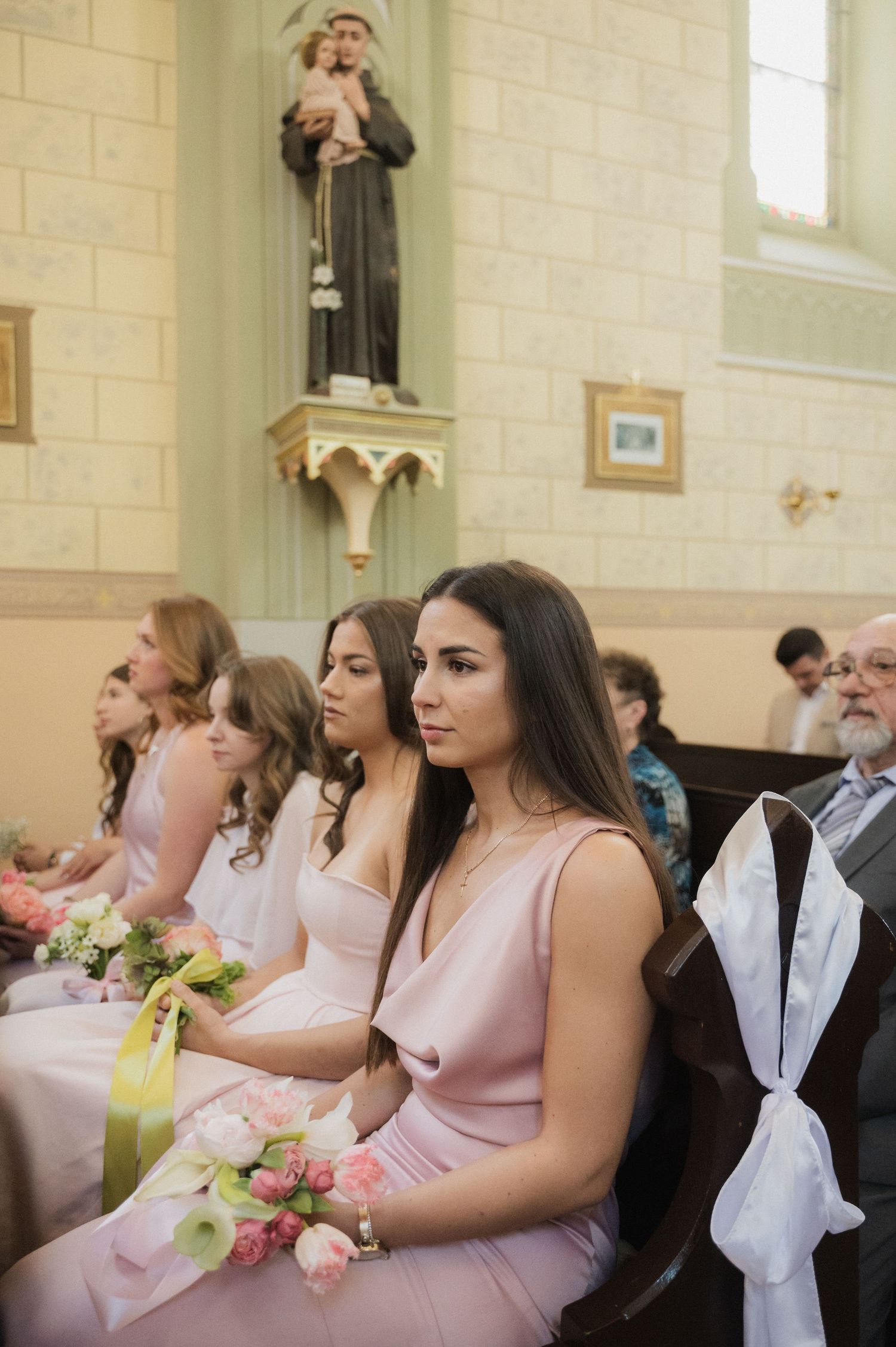 Bridesmaids in blush pink dresses seated in church pews during a wedding ceremony.