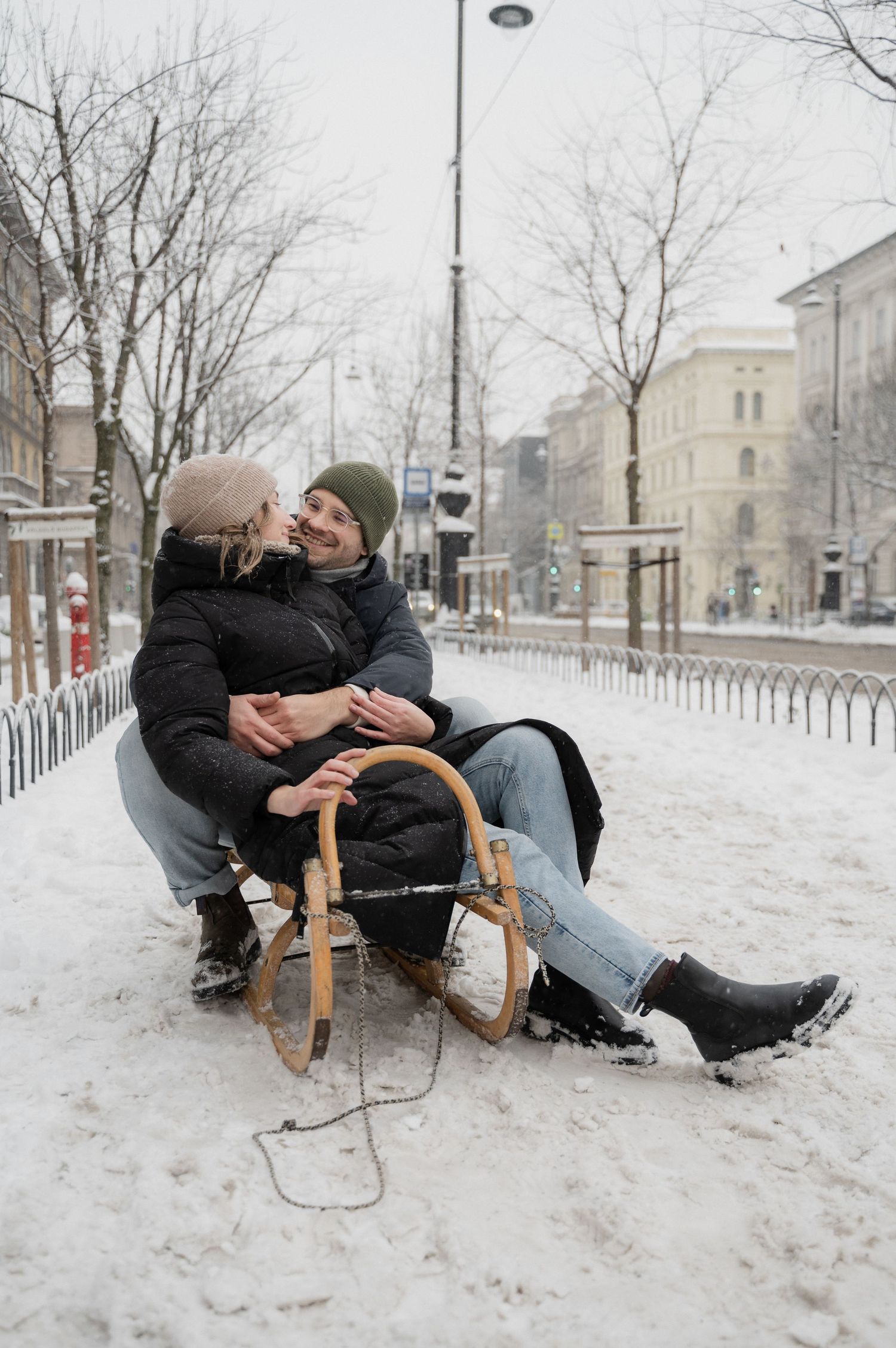 A couple sharing a romantic moment on a wooden sled in a snowy winter cityscape.