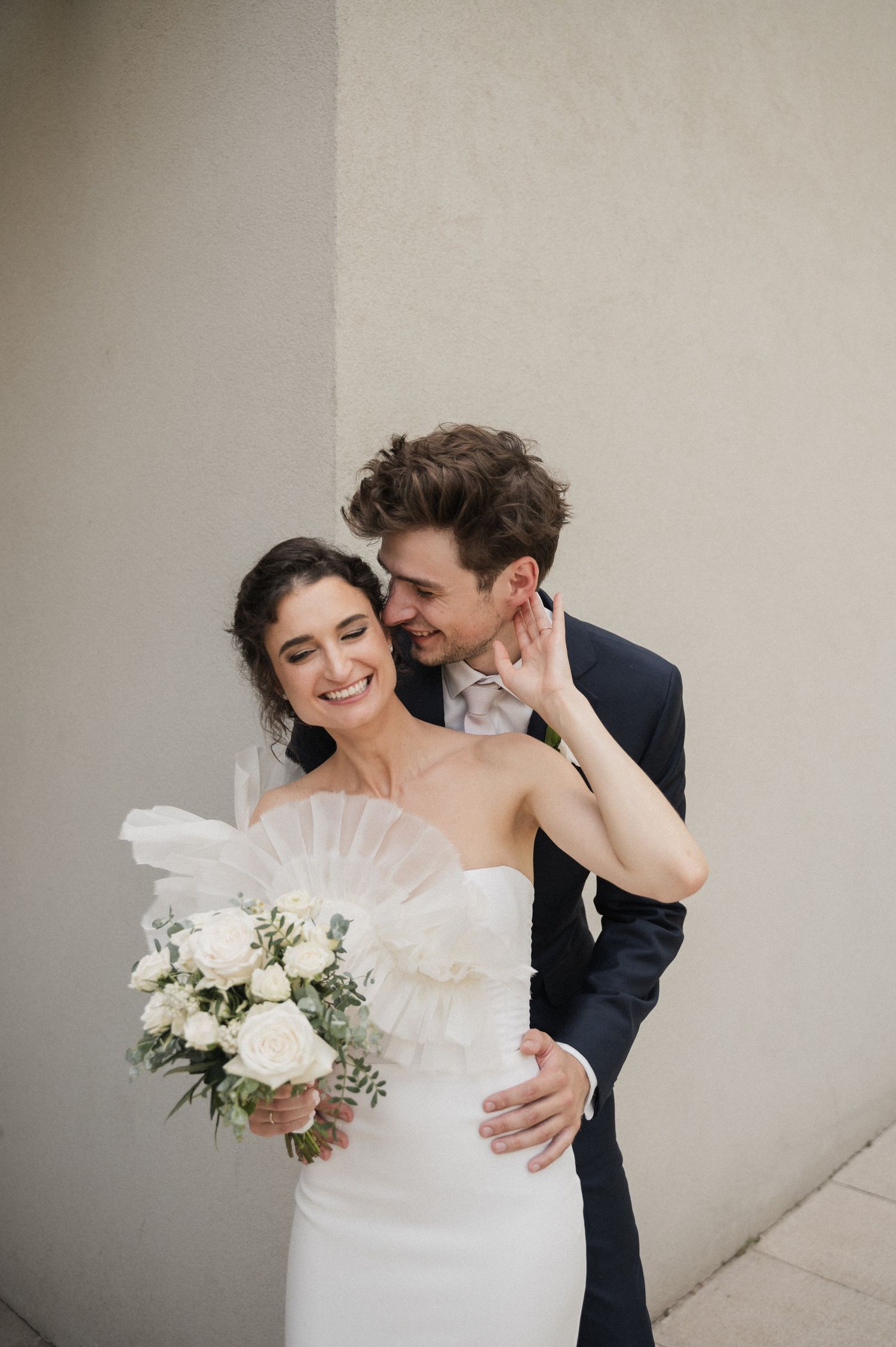 Romantic wedding couple embraces in an intimate moment against a neutral wall with white floral bouquet.