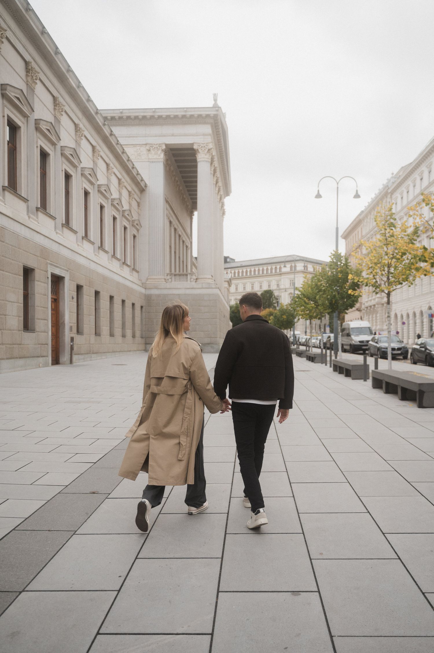 A couple walks hand in hand down a grand European street with classical architecture and street lamps.
