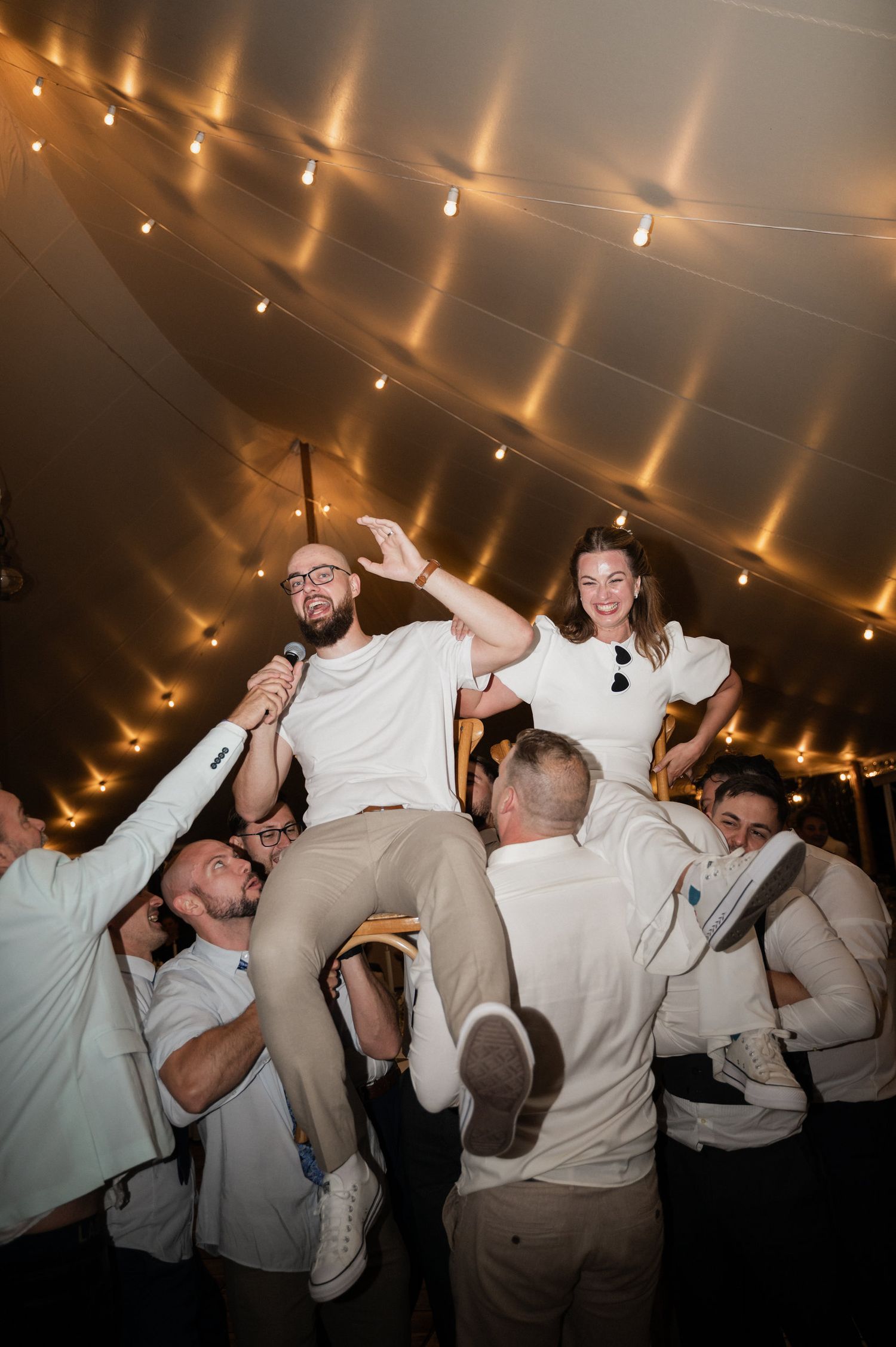 Wedding reception celebration with guests lifting the newlyweds in the air under string lights in a tent.