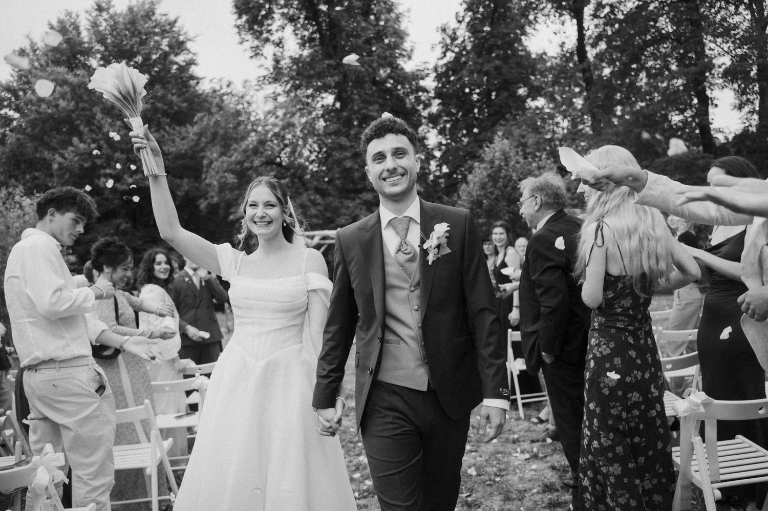 Joyful bride and groom walk through confetti shower outdoors surrounded by cheering wedding guests.
