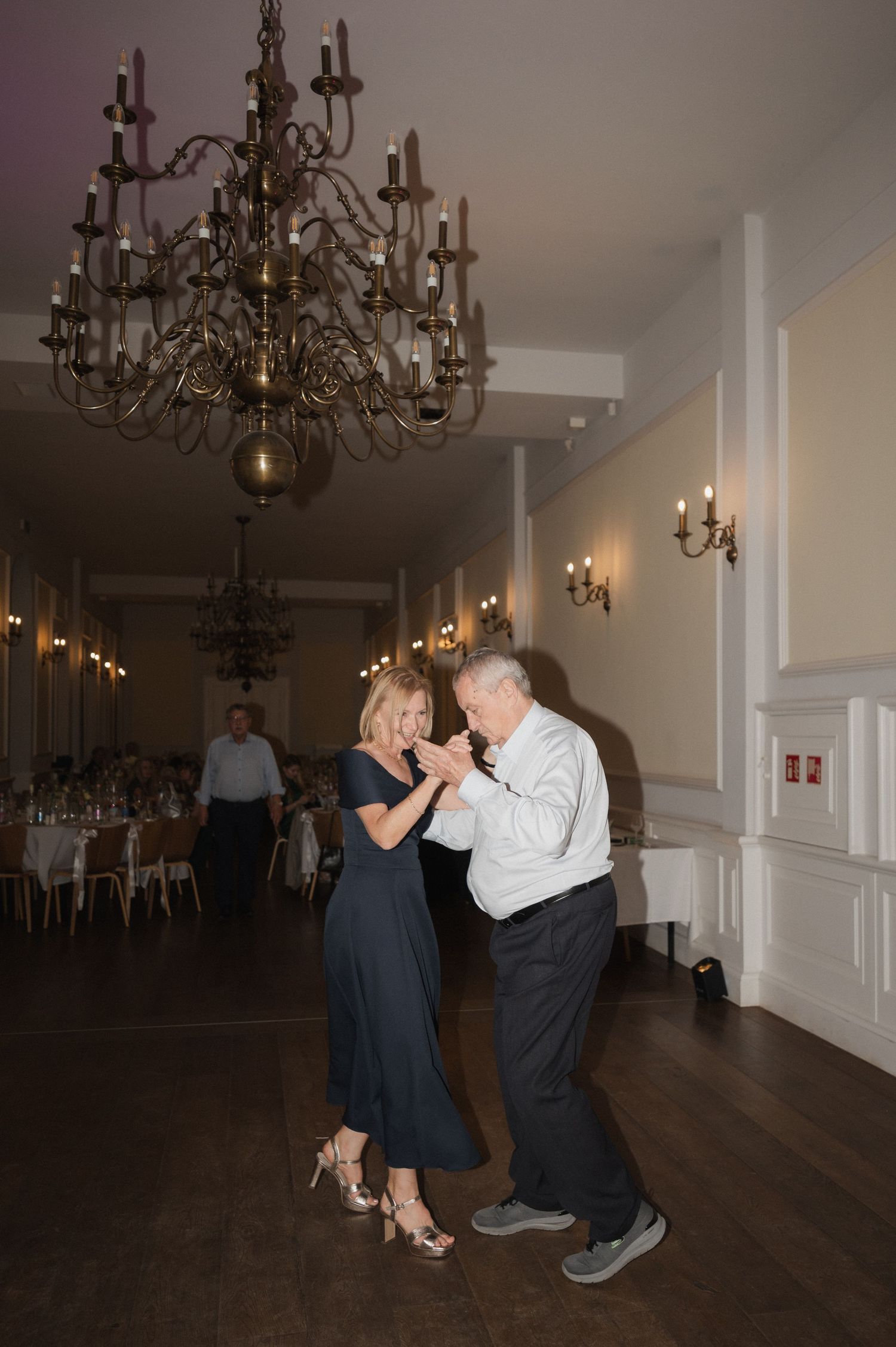 Couple dancing under ornate chandelier in grand ballroom during wedding reception with candlelit tables in background.