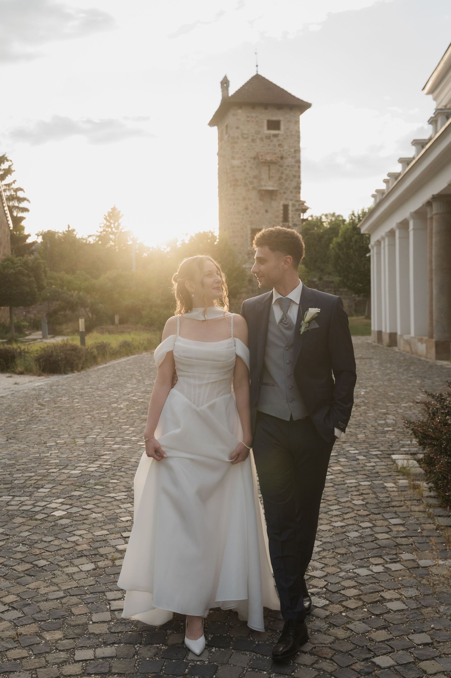 Bride and groom walk hand in hand at golden sunset with stone tower behind them.