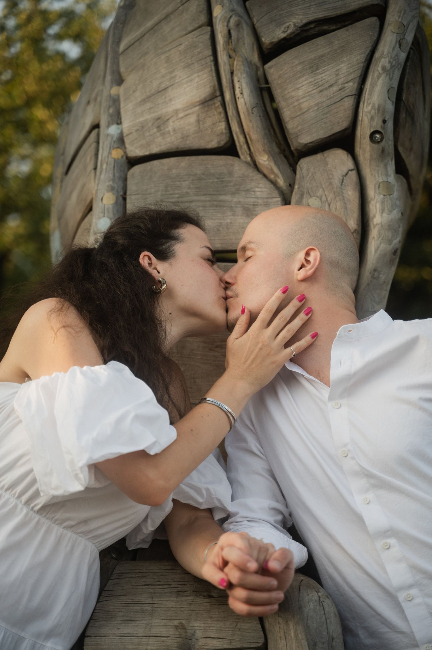 A romantic couple in white clothing share an intimate kiss against a backdrop of wooden sculptures.