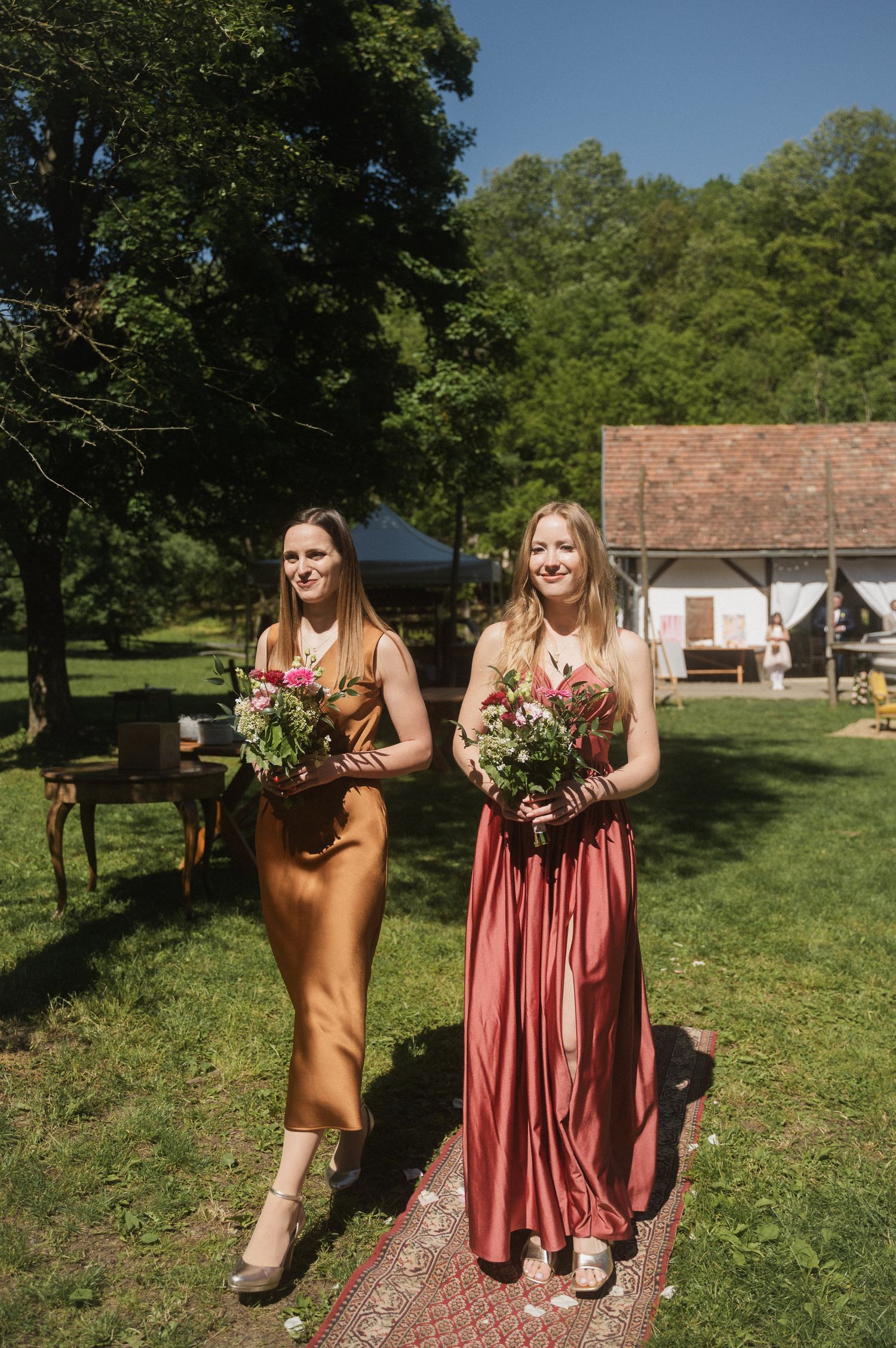 Two people in brown and pink dresses stand together on a grassy lawn near a rustic building on a sunny day.