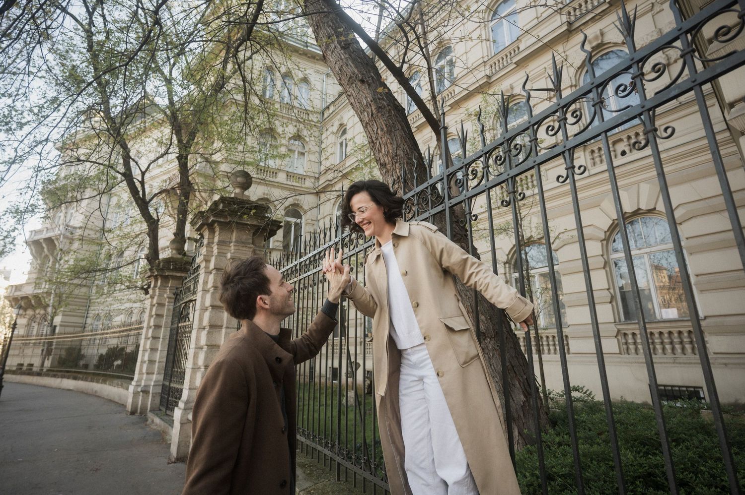 Two people share a tender moment by an ornate iron fence outside a historic building on a city street.