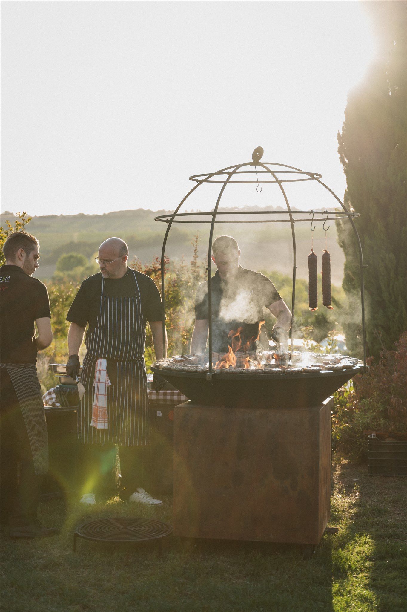Outdoor catering setup with chefs preparing food under gazebo structures at sunset.