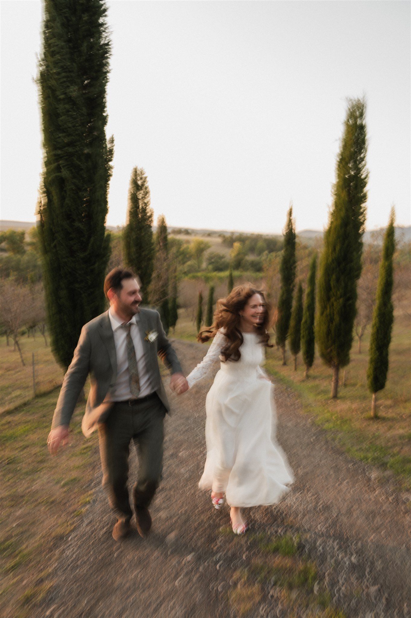 Joyful couple running through cypress-lined path in Italian countryside setting.