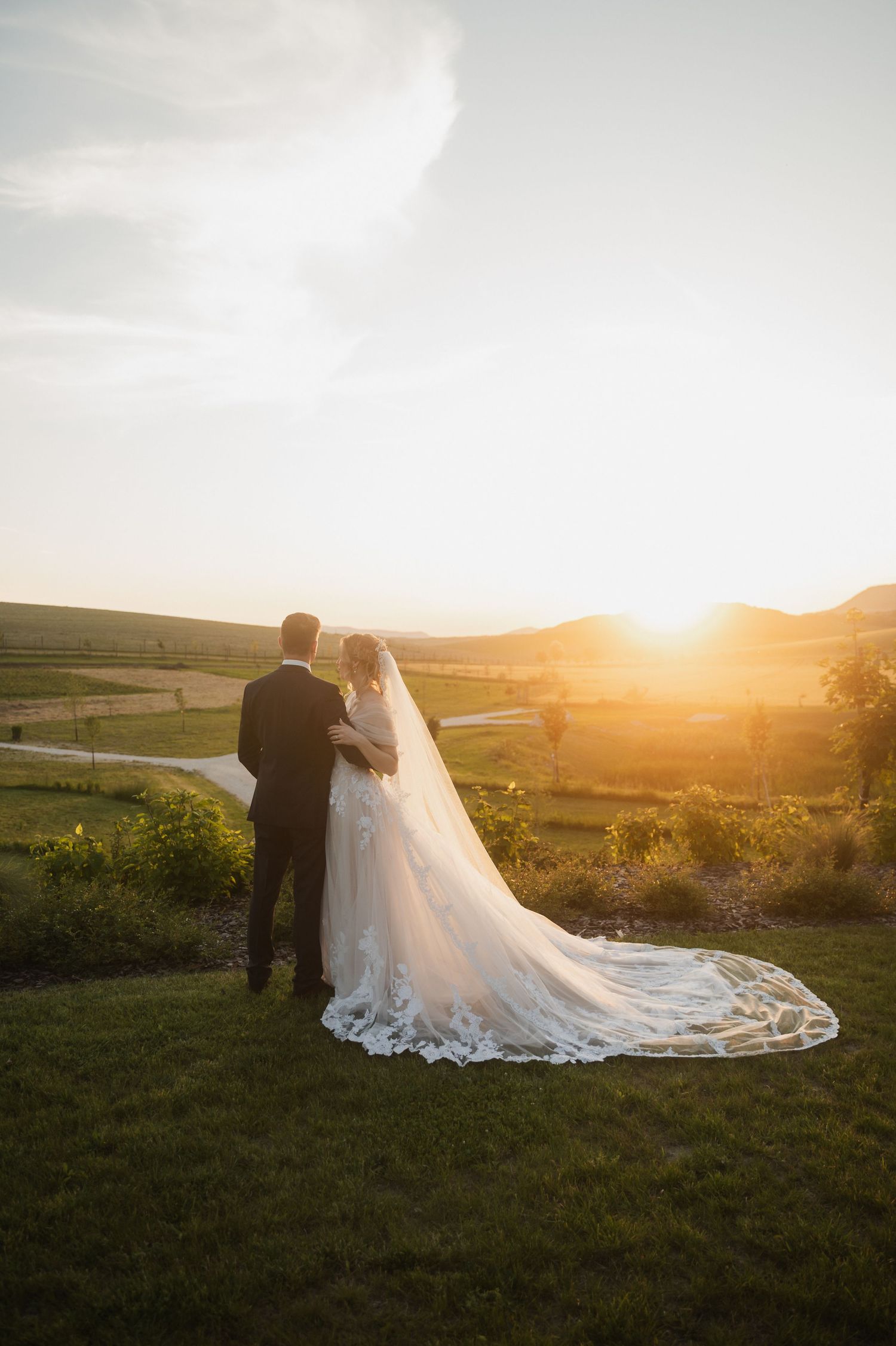 Newlywed couple embraces during golden sunset in rolling countryside with long flowing wedding veil trailing behind.