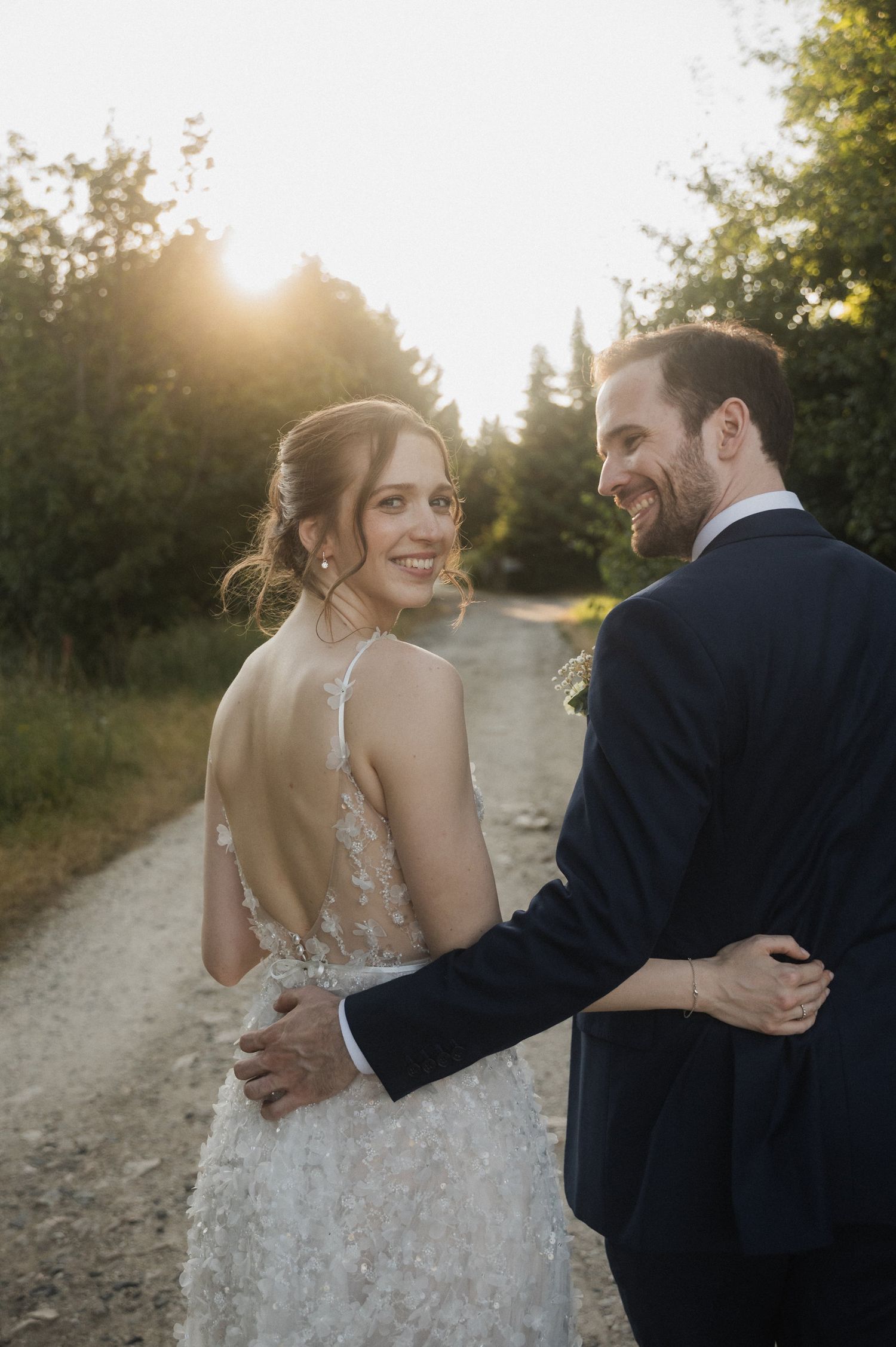 A romantic wedding couple embraces on a sunlit dirt path surrounded by trees during golden hour.