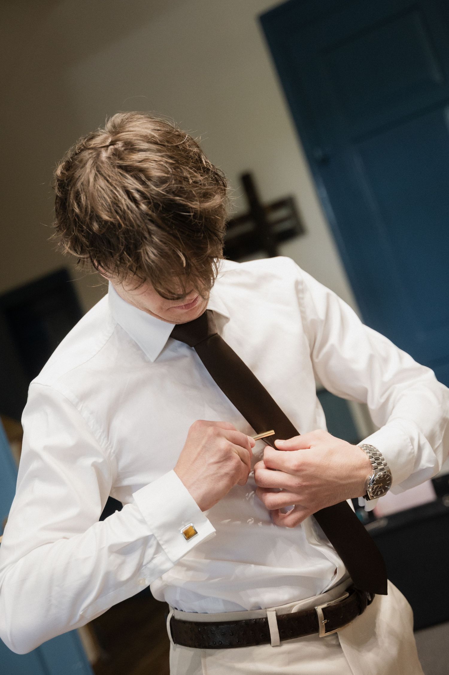 Groom in white dress shirt and dark tie fastening cufflinks while getting ready for his wedding day.