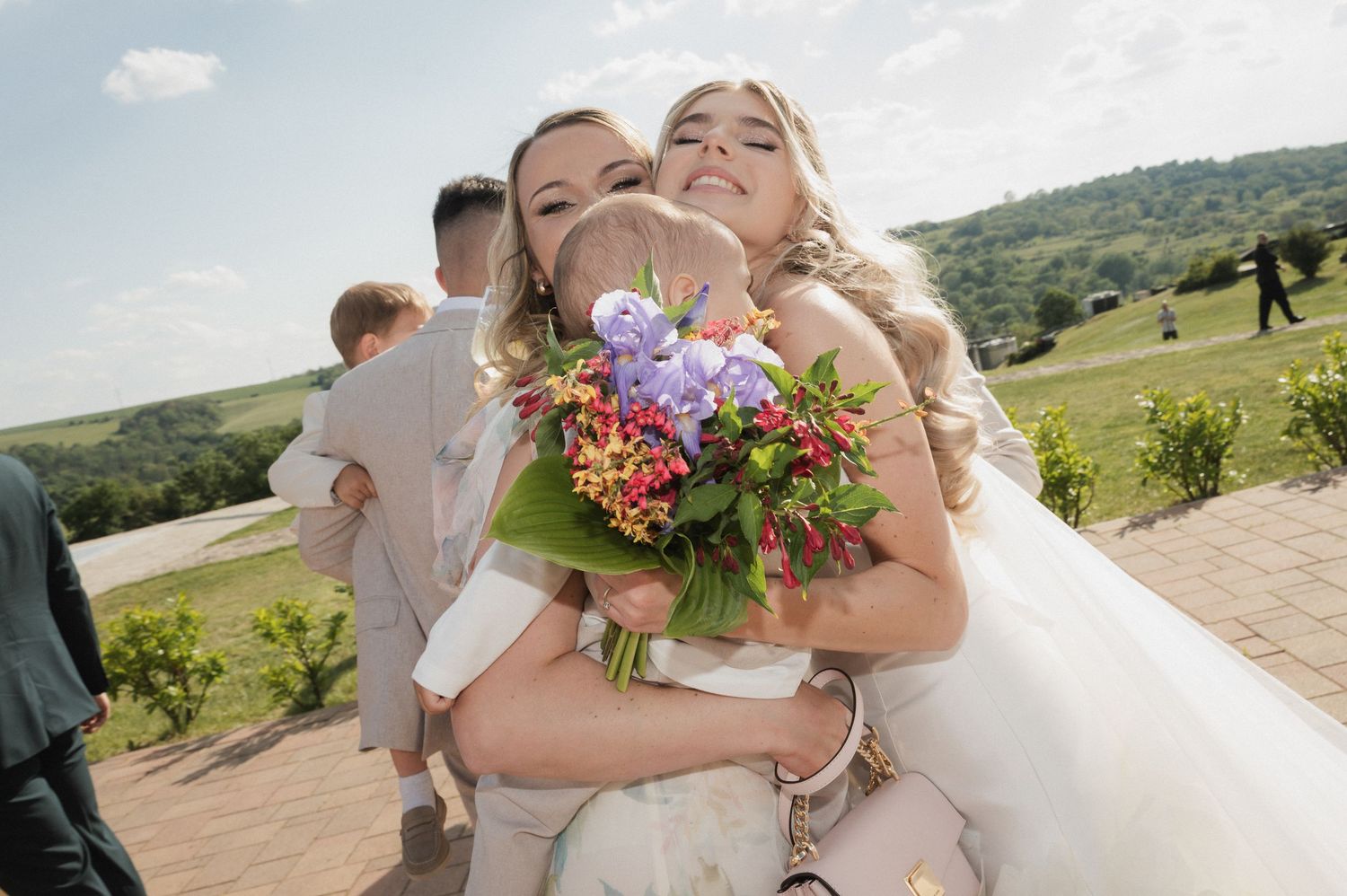 Bride hugged by bridesmaids while holding colorful bouquet at outdoor vineyard wedding.