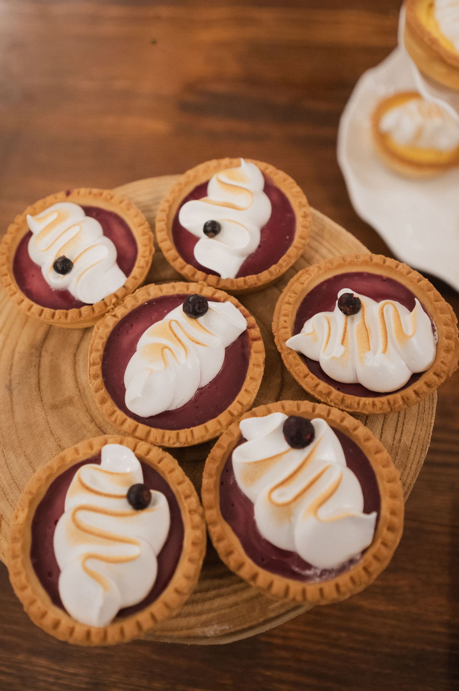 Five berry tart desserts topped with swirled meringue and blueberries displayed on a wooden board.