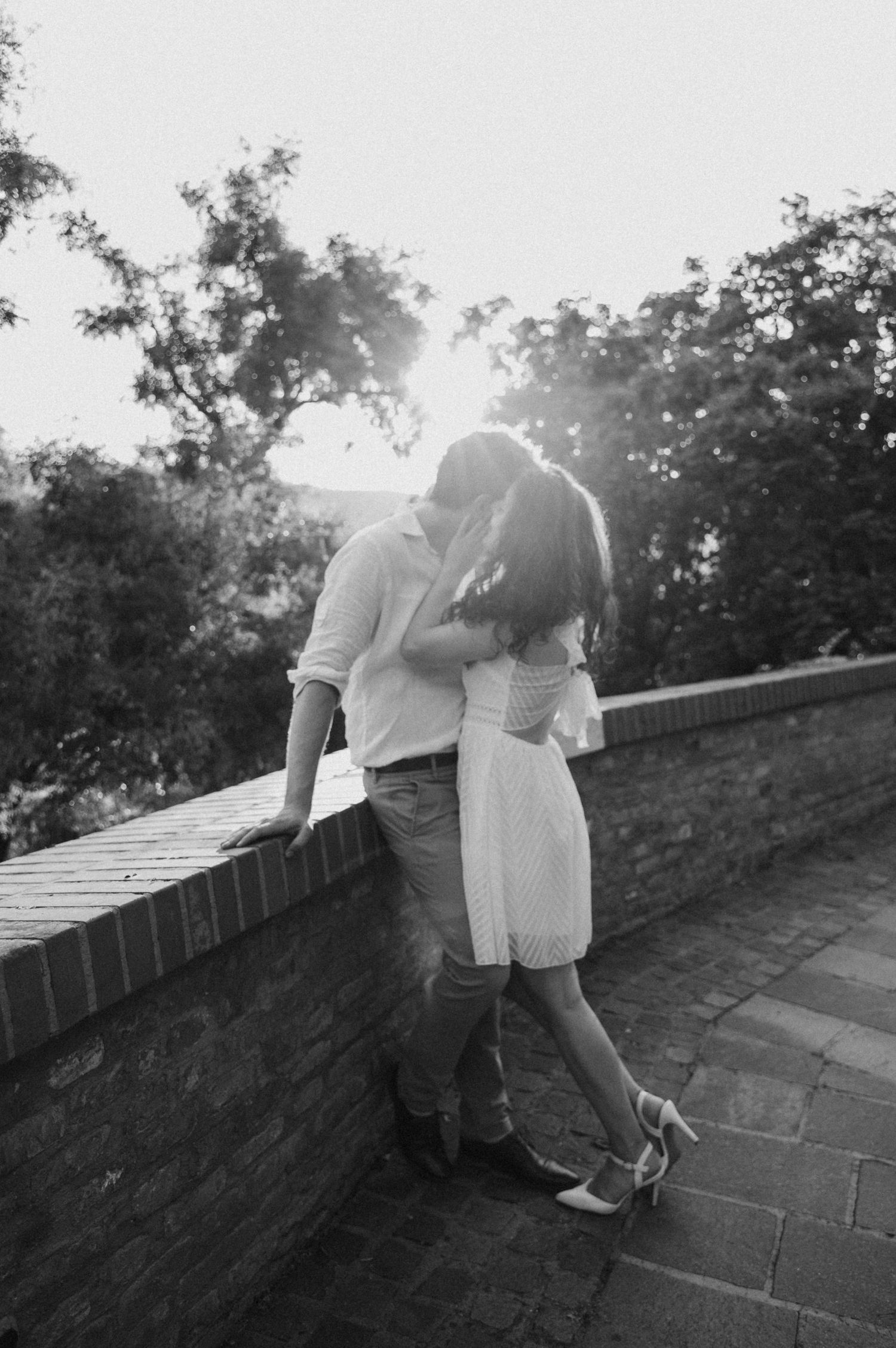 A romantic black and white photograph of a couple embracing against a brick wall at sunset.