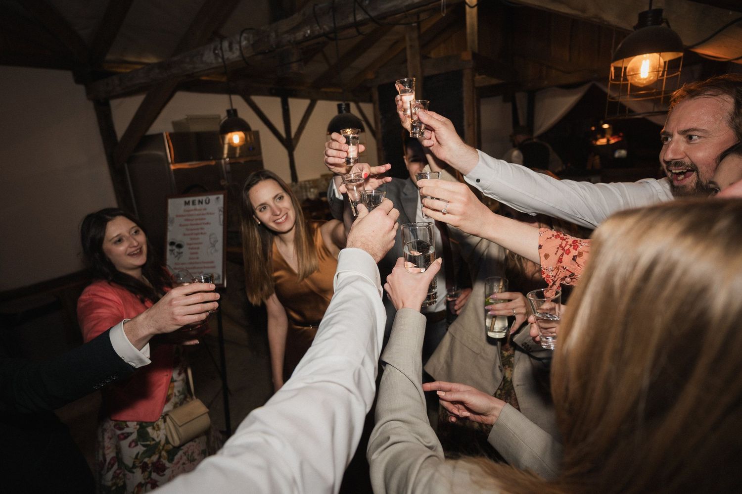 Friends raising glasses in a celebratory toast at a rustic indoor venue with wooden beams overhead.