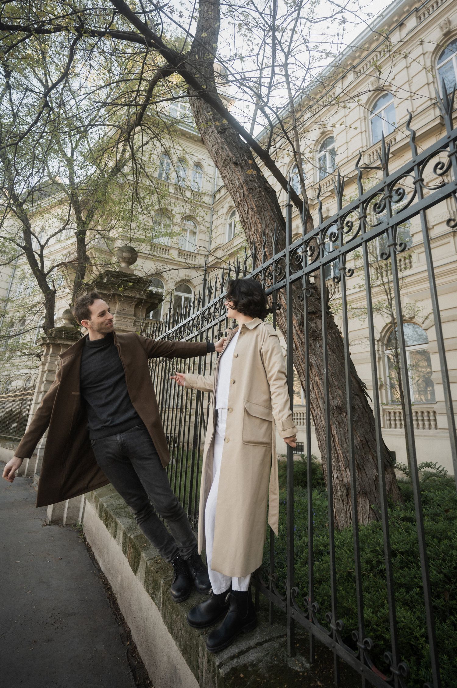 A person in dark clothing climbs over an ornate iron fence outside a historic building in an urban setting.